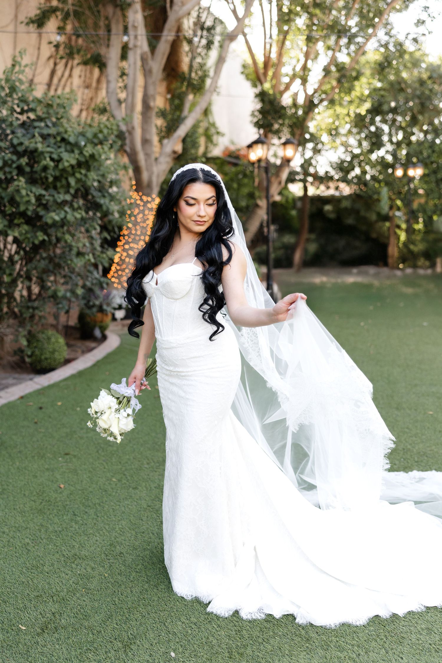 A bride in a white lace wedding dress poses gracefully on a manicured lawn during an outdoor photo session.