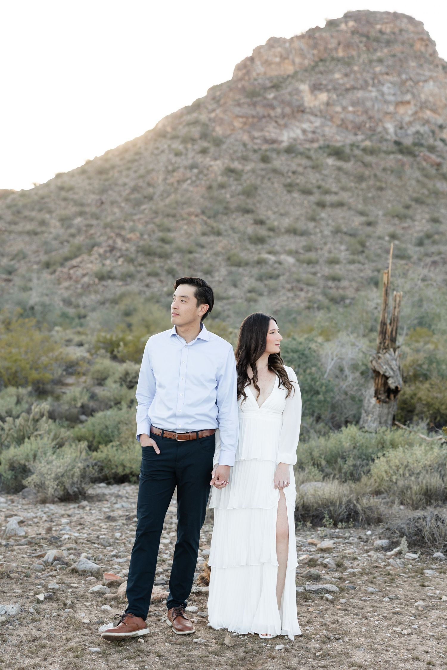 A couple in formal attire poses against a desert mountain backdrop with sagebrush and rocky terrain.