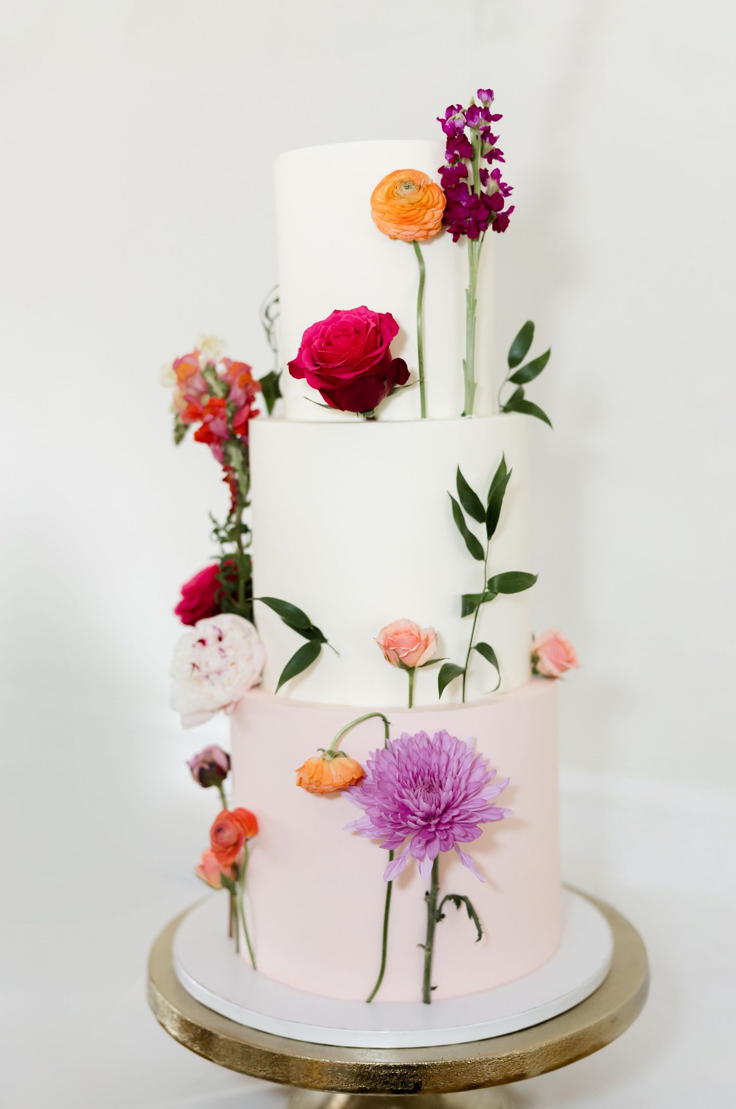 Elegant two-tier white wedding cake decorated with fresh colorful flowers on a gold base.