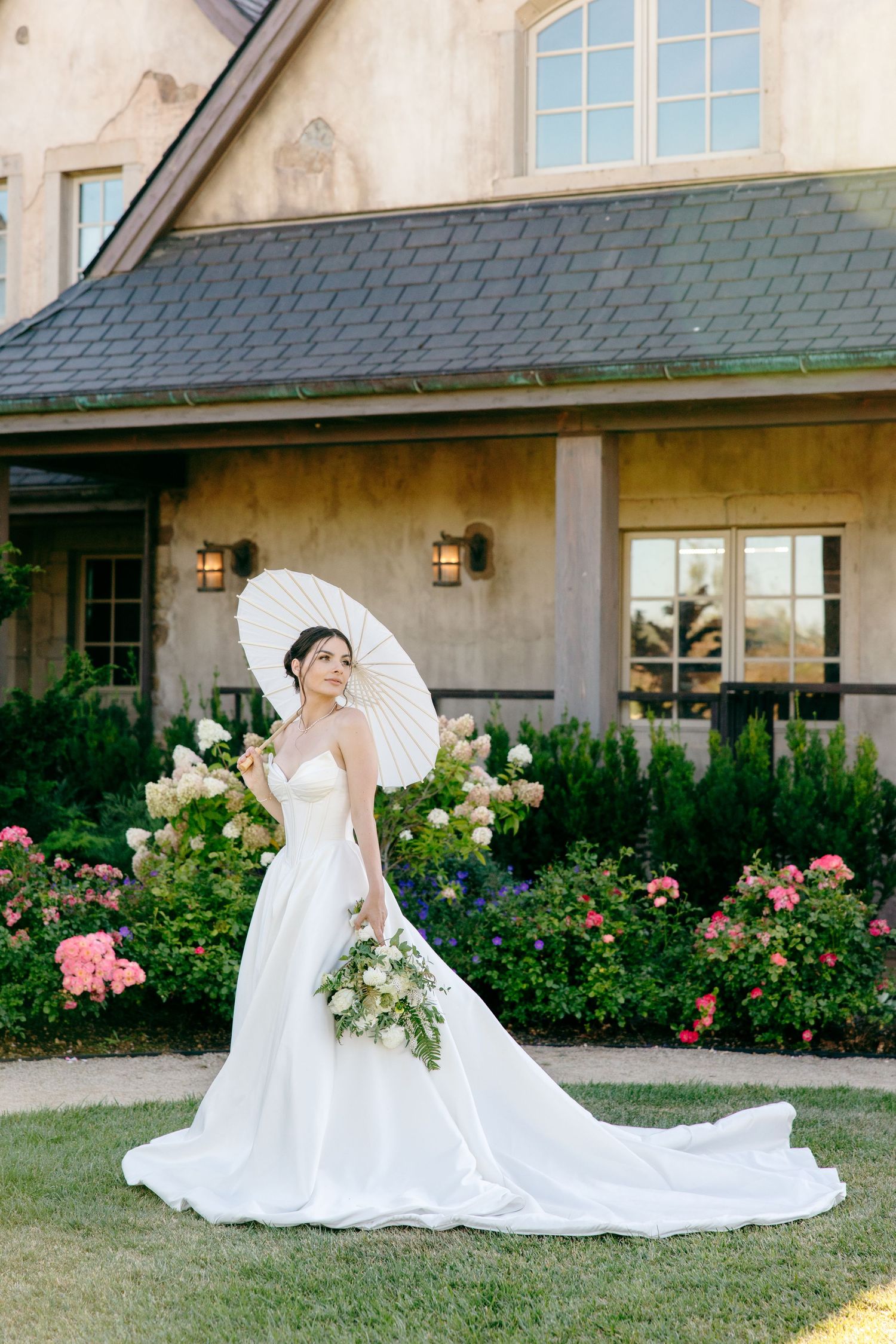 Bride in flowing white wedding gown with long train poses gracefully on manicured lawn beside rose garden.