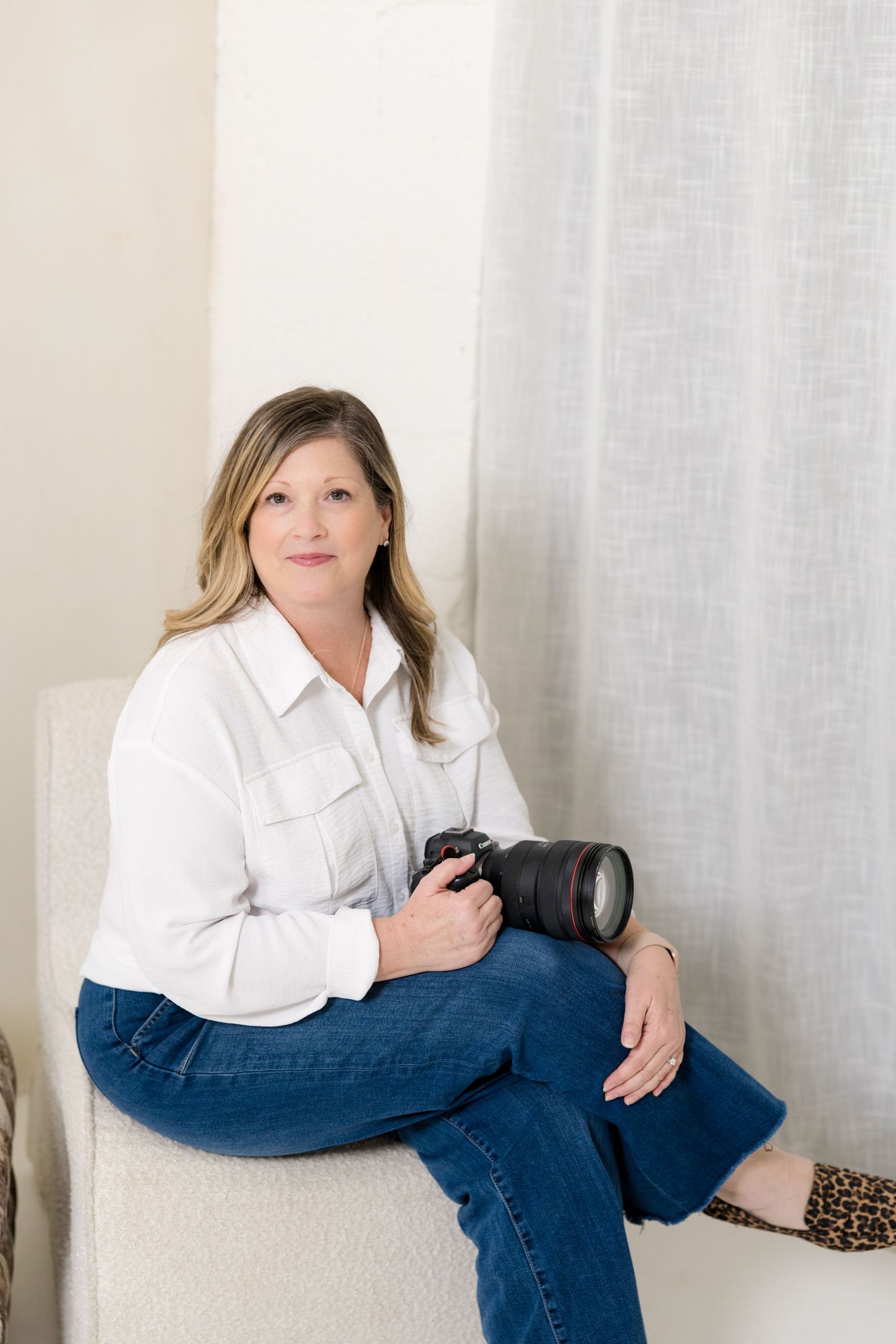 Professional photographer in white button-down shirt and jeans poses with camera in studio setting.