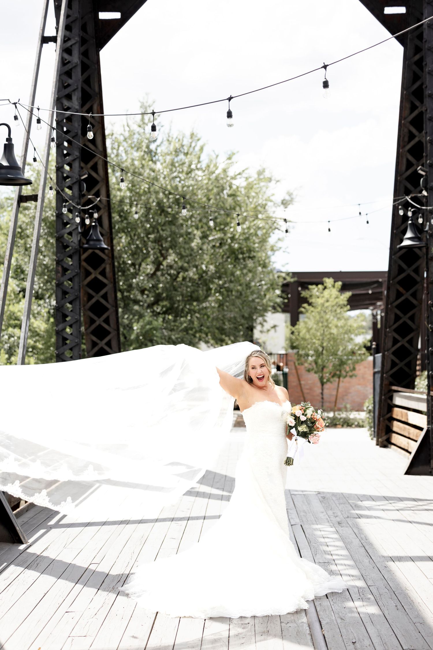 Joyful bride in white gown with flowing veil holds bouquet on wooden deck with string lights and iron structure.