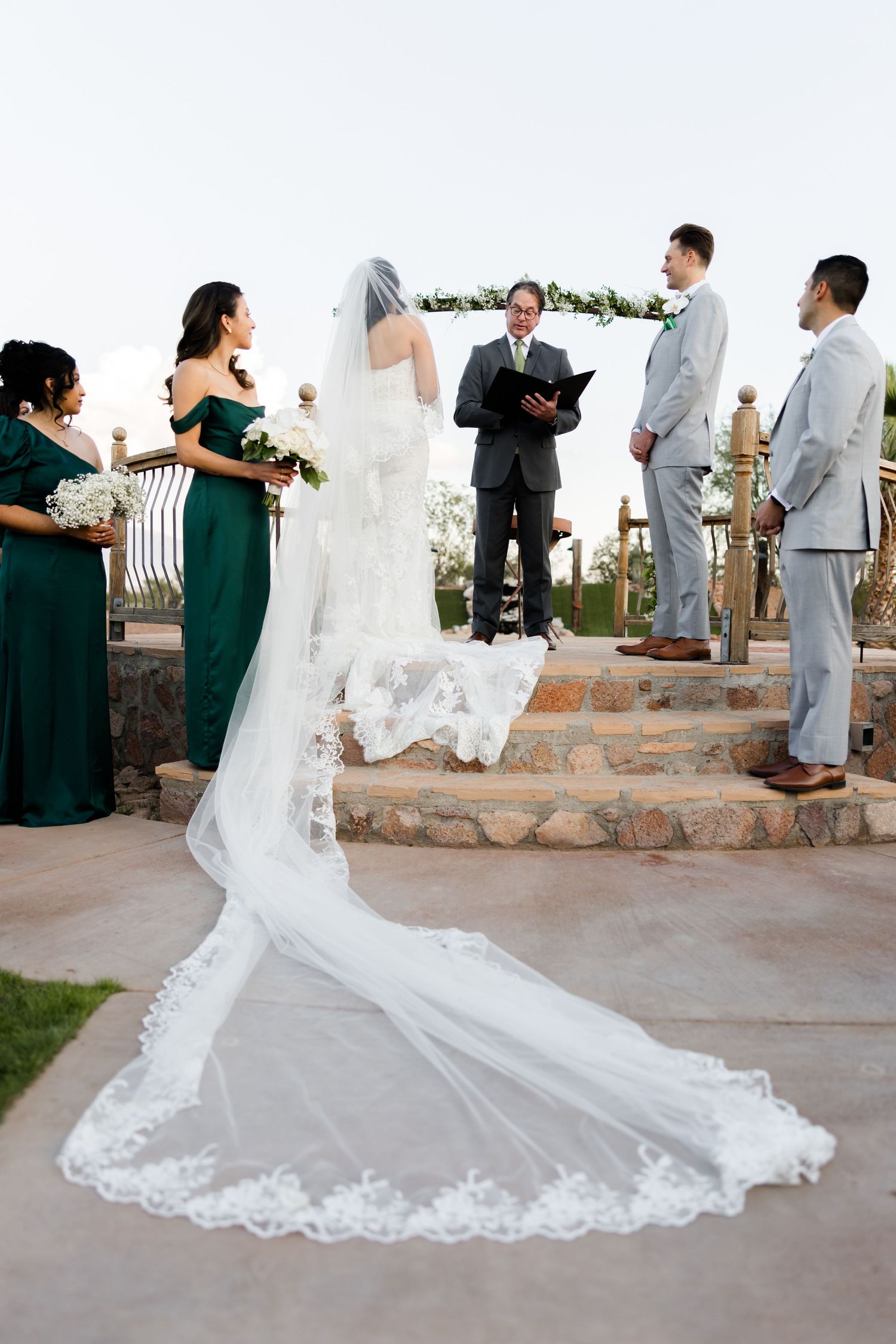 Bride in white gown with long lace veil stands at outdoor wedding altar with bridesmaids in green dresses.