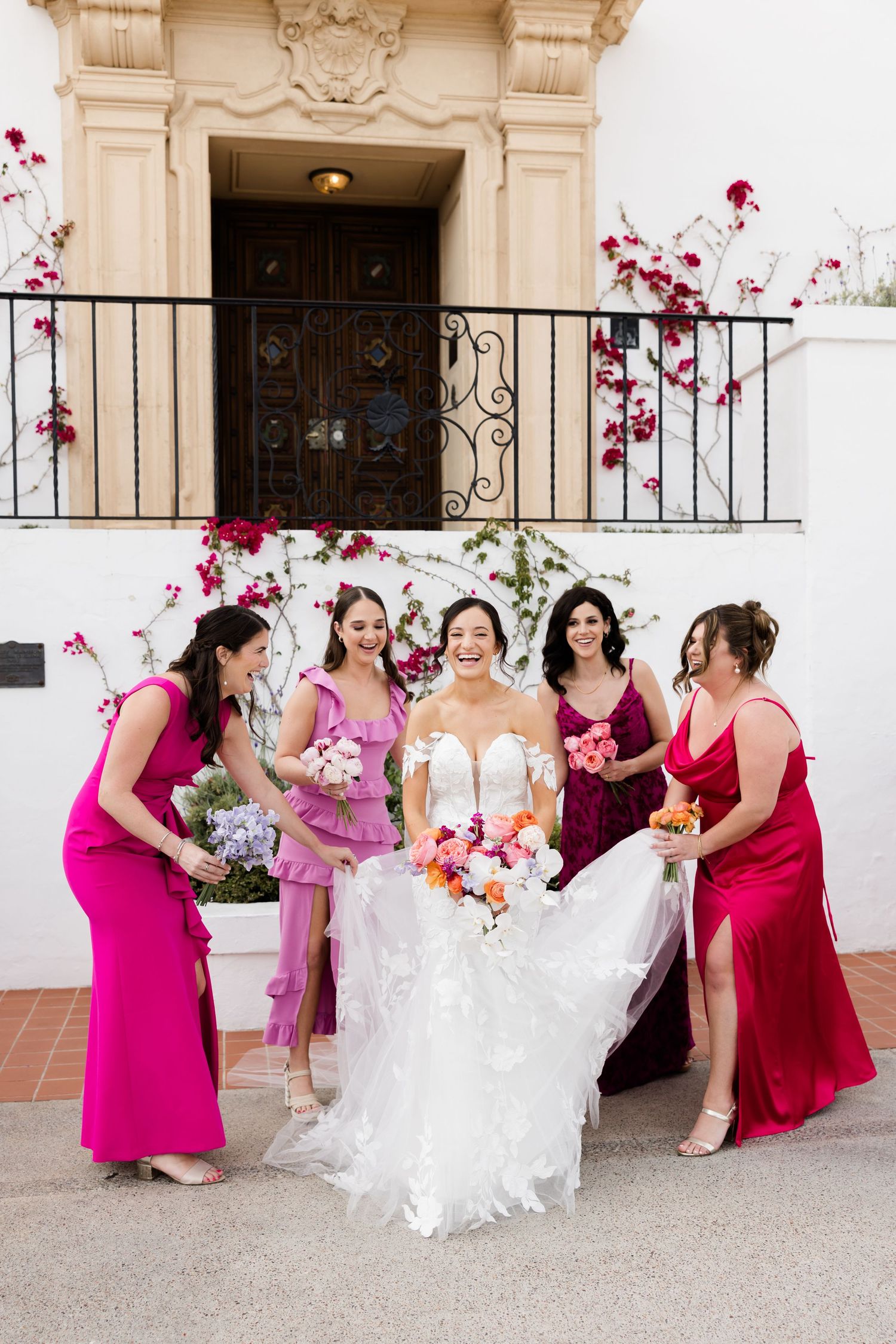 Bridesmaids in vibrant magenta dresses helping arrange bride's gown against white colonial backdrop.