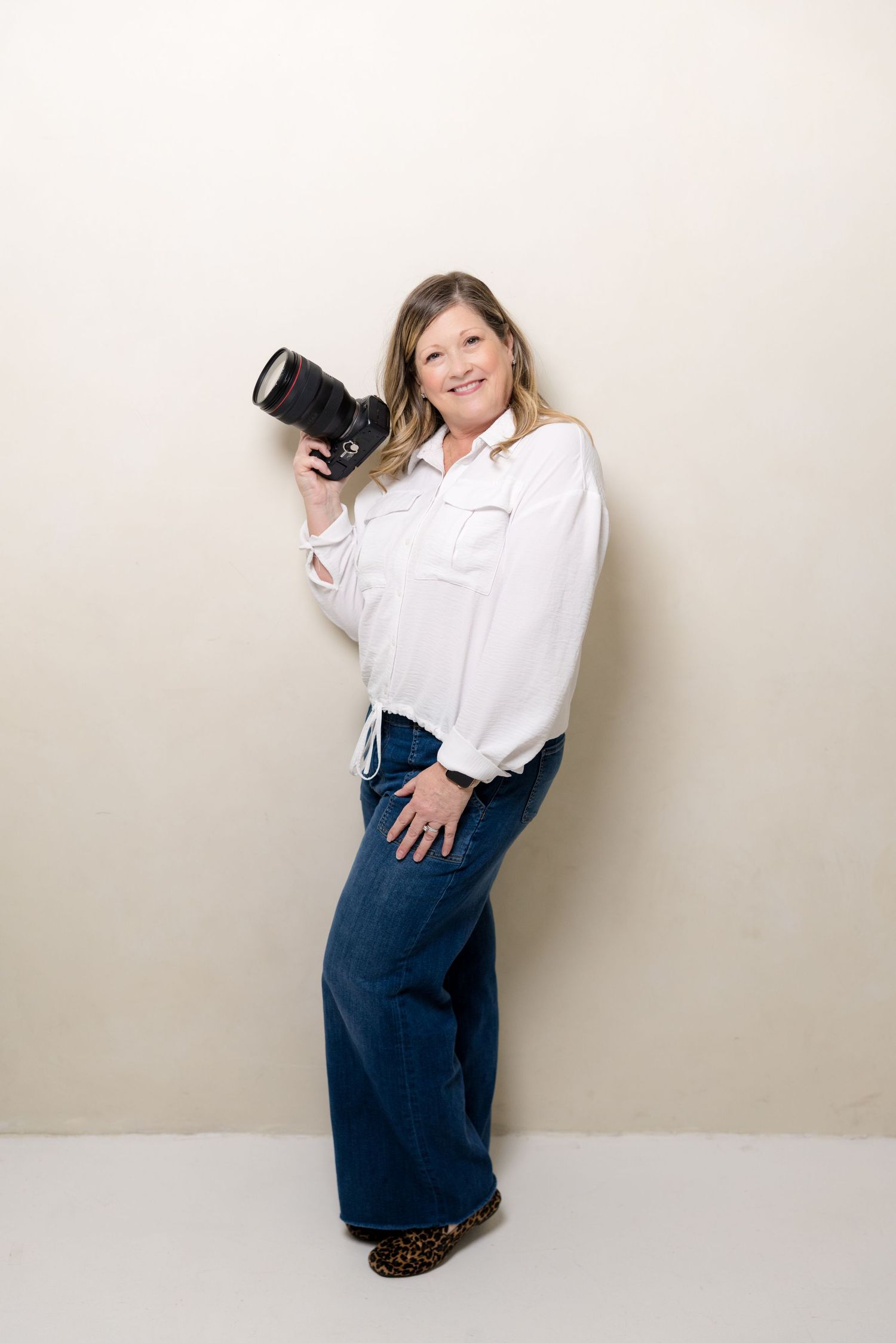 Professional photographer in white button-down shirt and navy skirt poses with camera against neutral background.