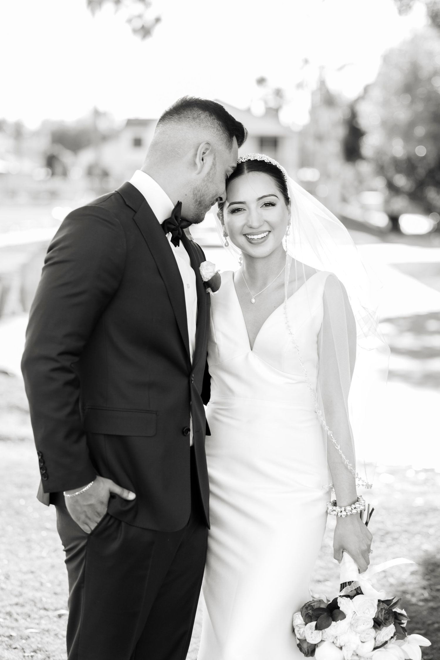 Black and white wedding portrait of smiling bride in veil holding bouquet leaning close to groom in tuxedo outdoors.