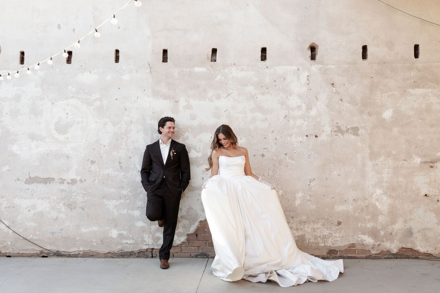 Bride in white ball gown and groom in black suit pose against rustic concrete wall at elegant wedding venue.