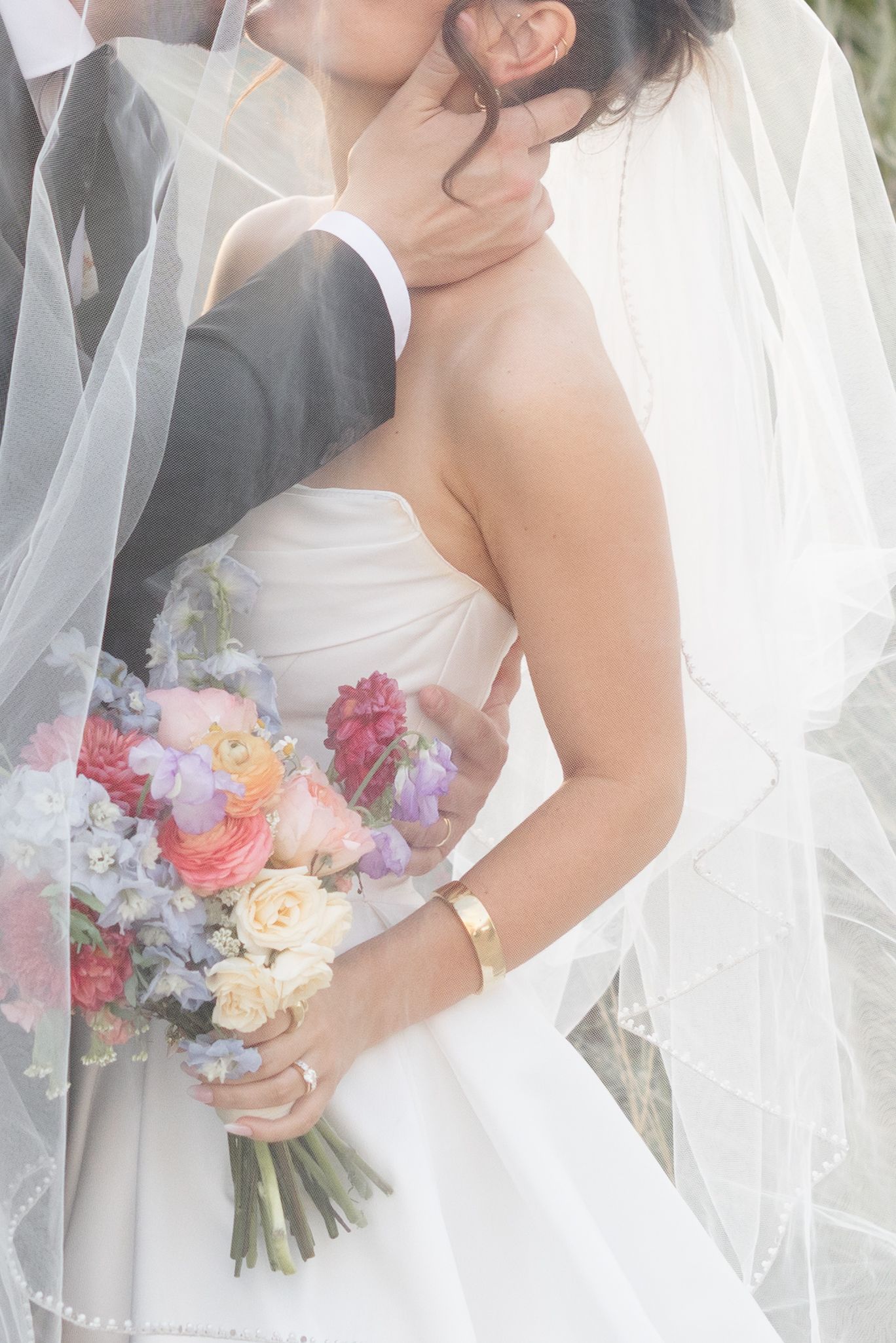 Bride in strapless white gown holds colorful bouquet while groom embraces her, both partially covered by flowing veil.
