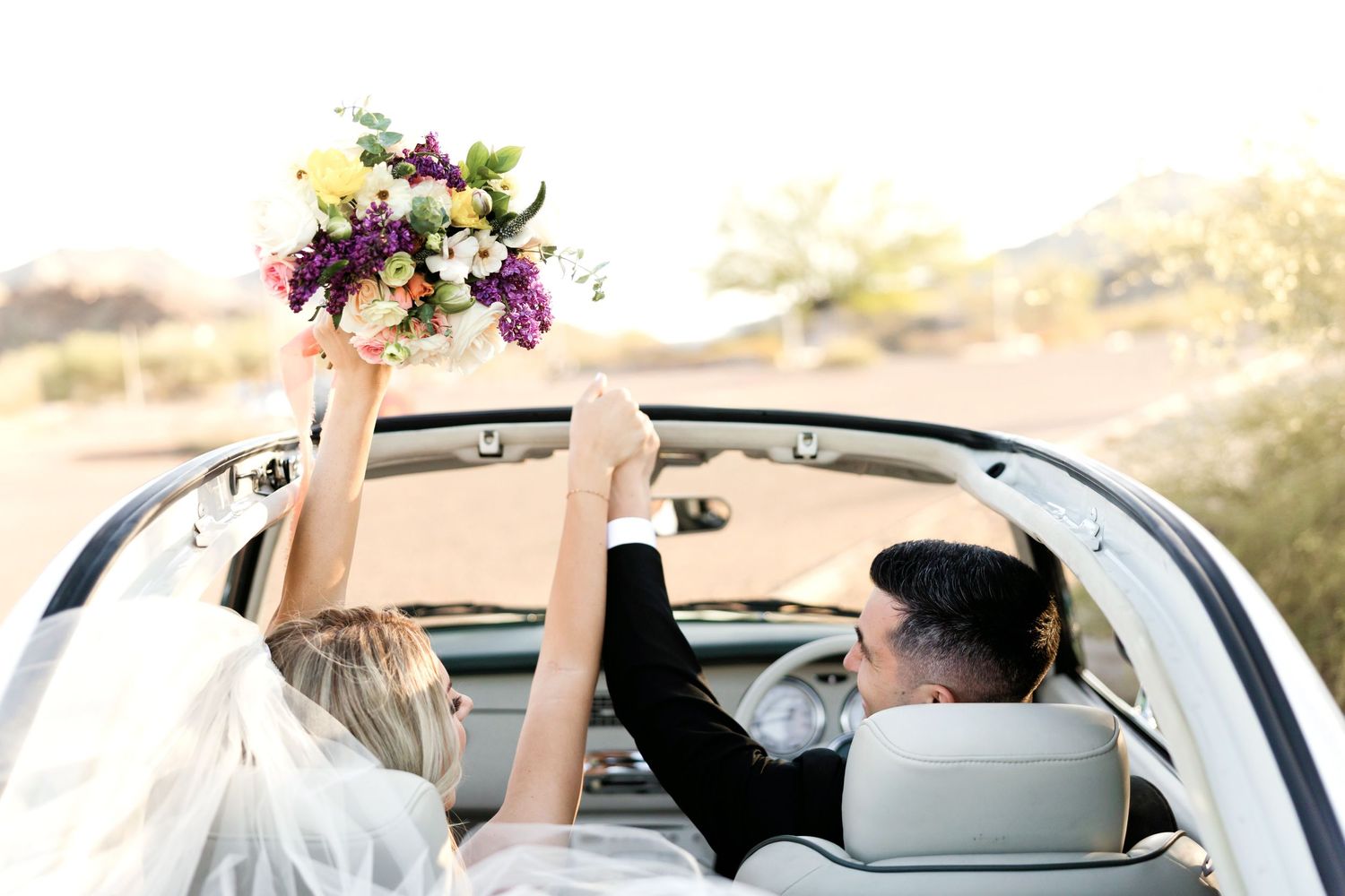 A couple in a convertible car celebrating their wedding day with a raised bouquet of flowers.