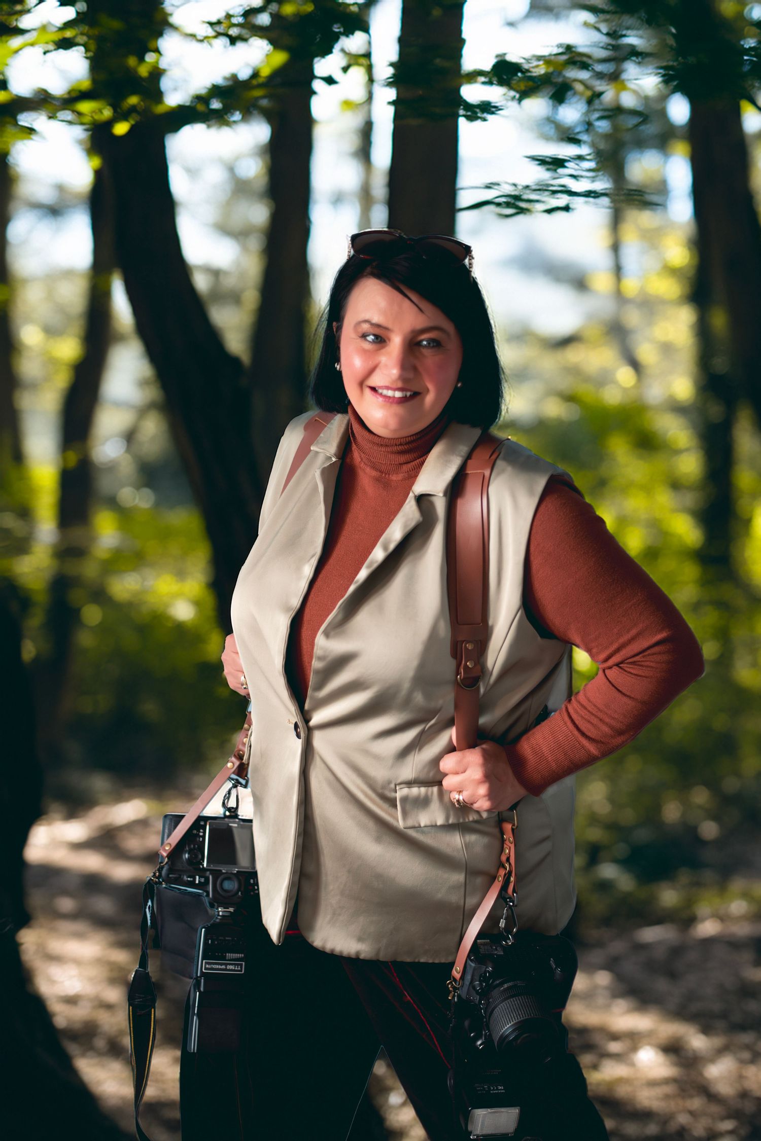 A person in a beige vest and brown shirt poses playfully in a sunlit forest setting.