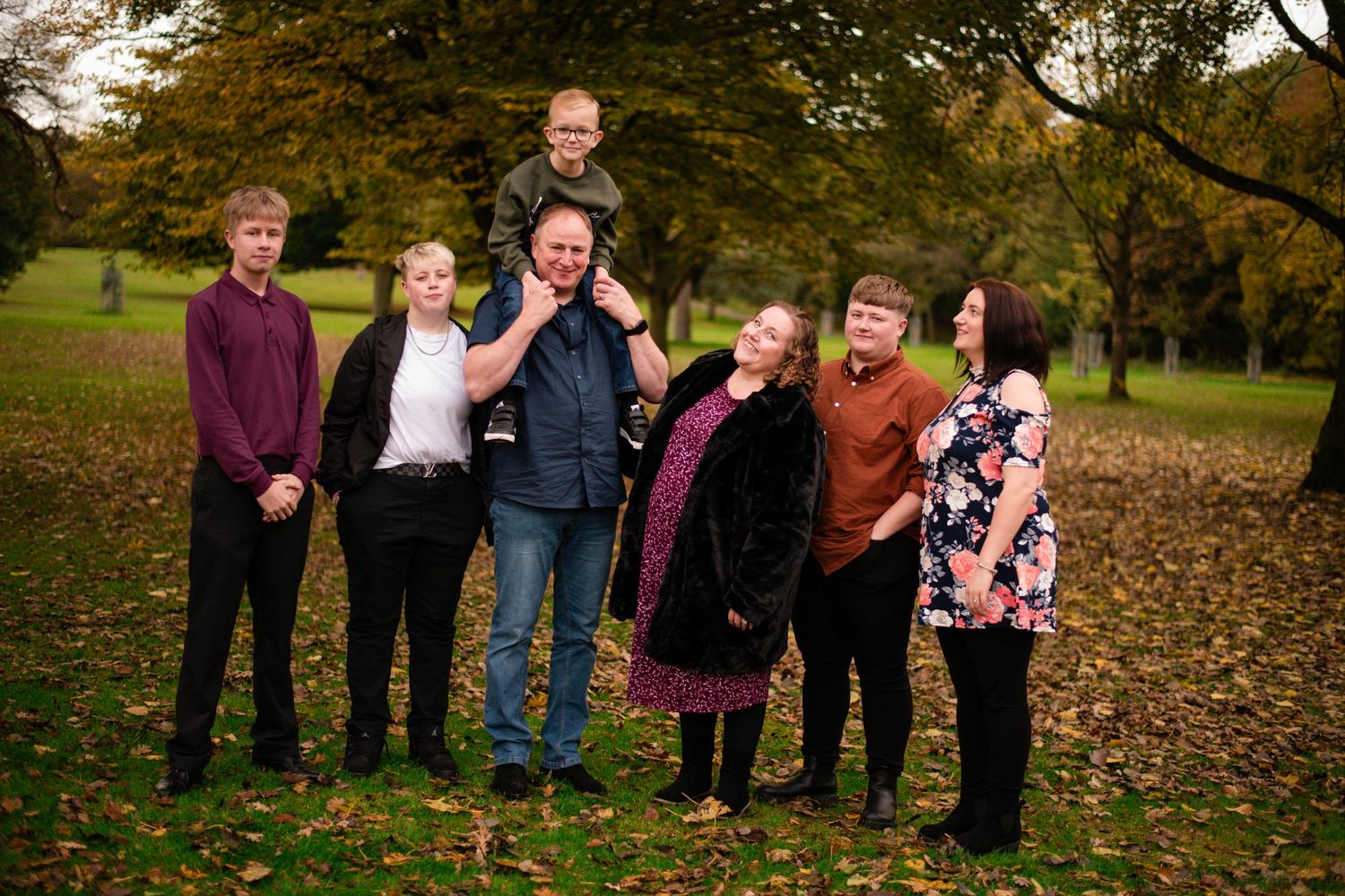 Family group photo taken outdoors in a fall setting with autumn leaves on the ground.