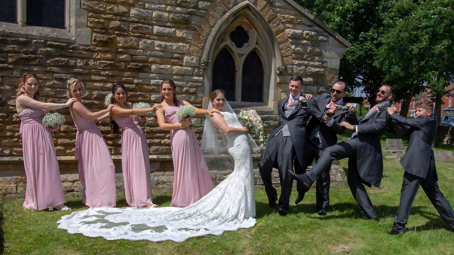 Wedding party poses playfully on church lawn with bridesmaids in pink and groomsmen in dark suits.