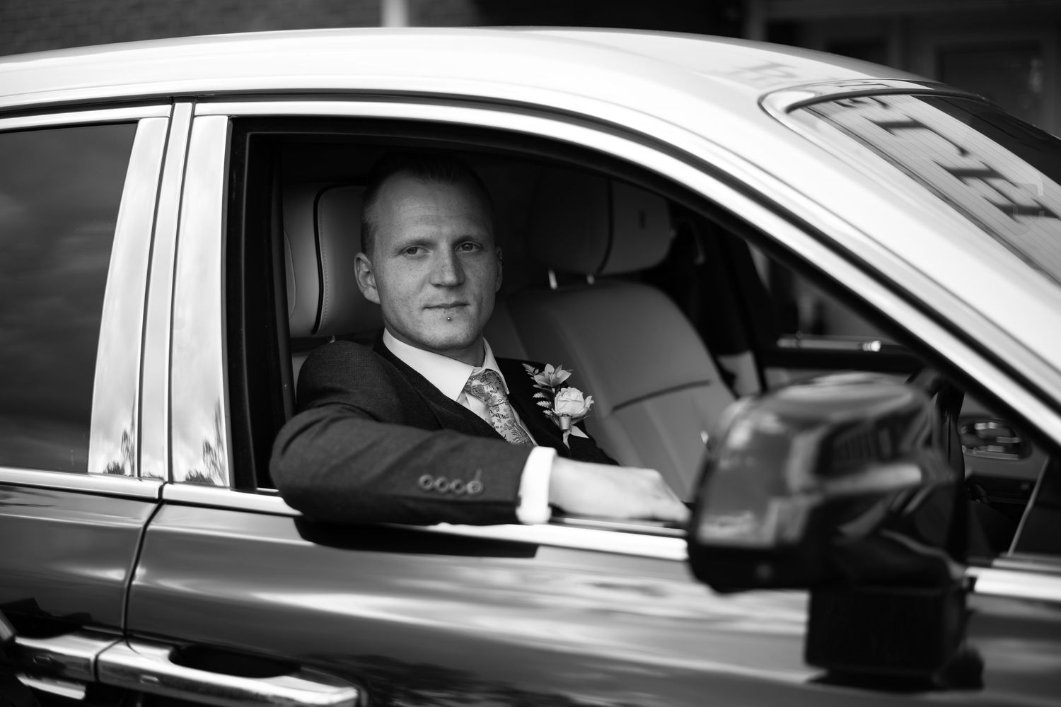 Black and white photo of a groom in a suit with boutonniere sitting in a luxury car on his wedding day.