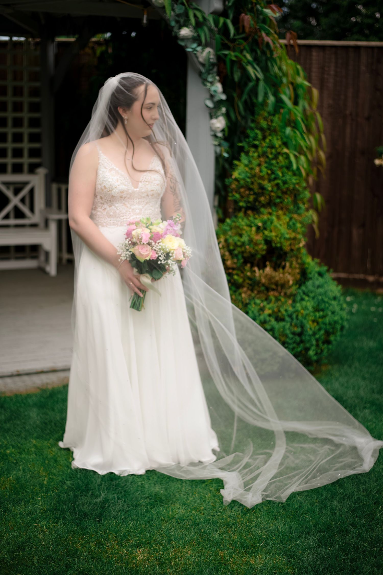 Bride in white lace gown with flowing veil holding pink bouquet standing on green lawn in garden setting.