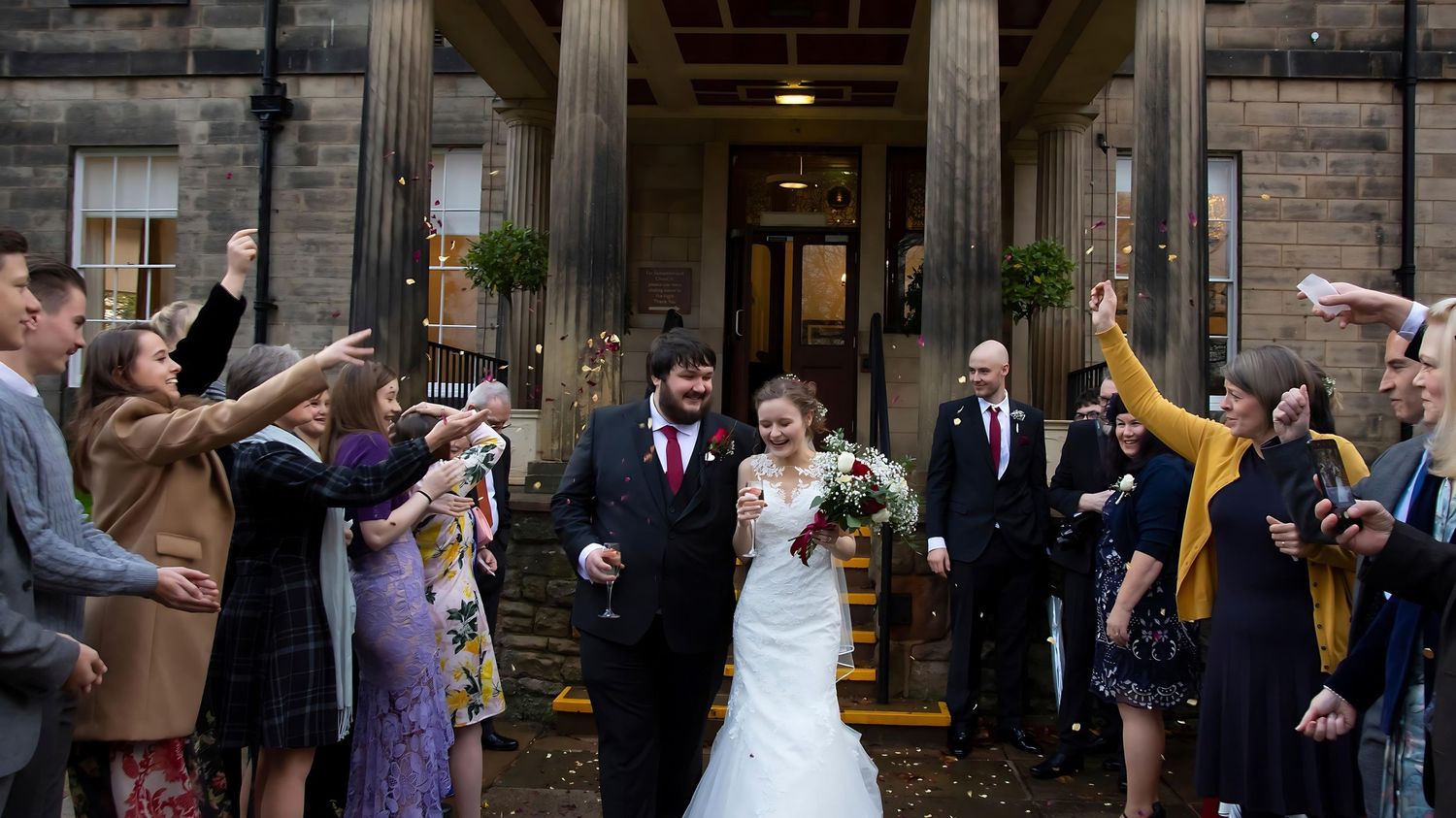 Wedding party and guests throw confetti outside historic stone pillared building.