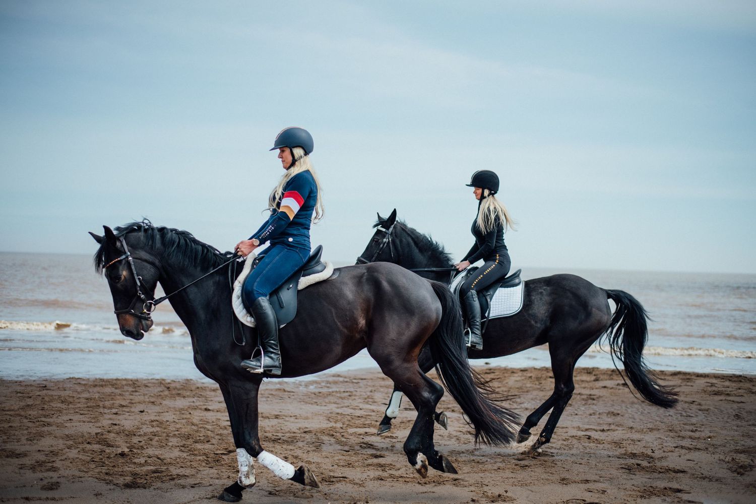 Two equestrians ride black horses in tandem on a sandy beach.