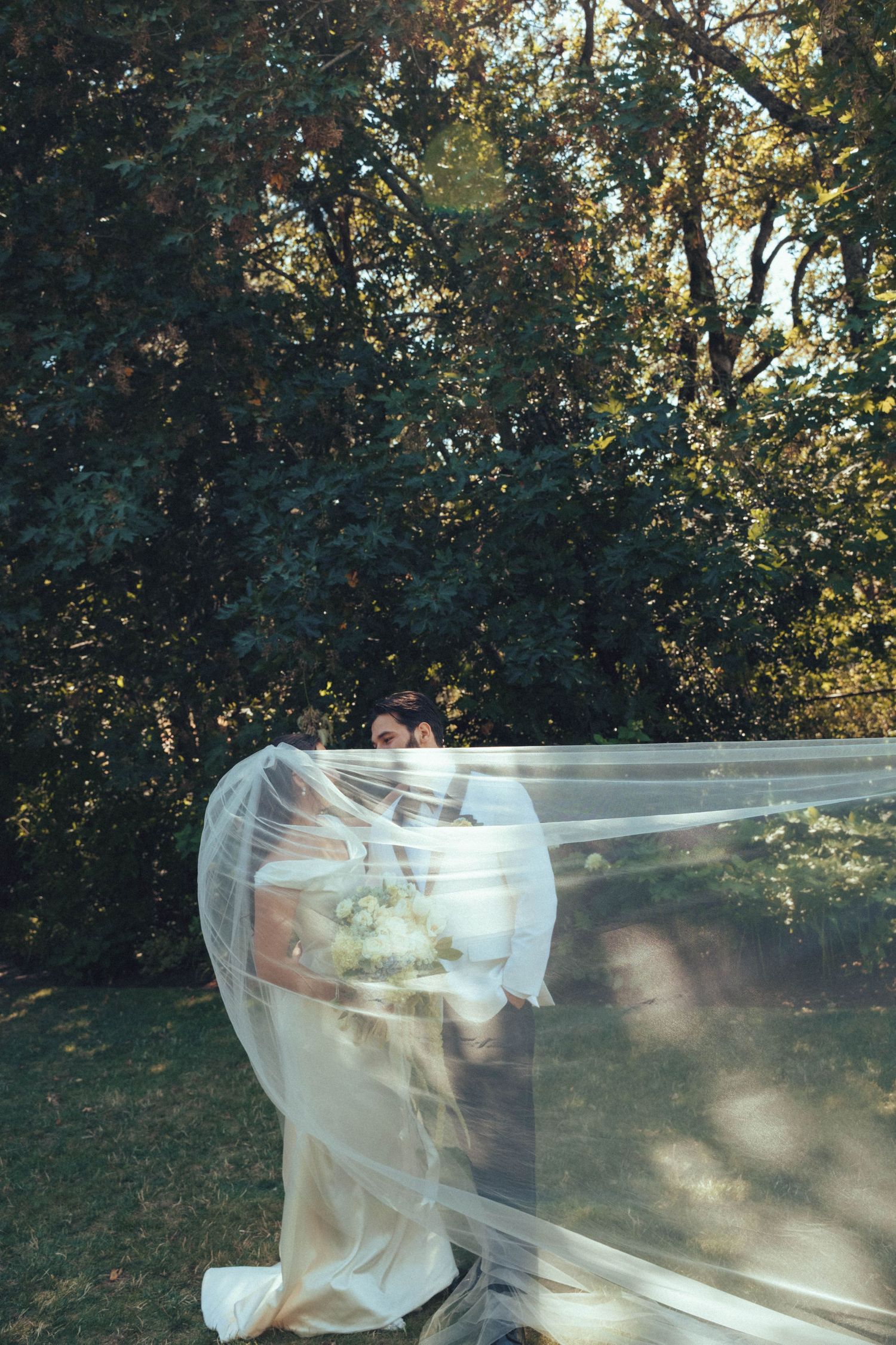 Bride and groom embrace outdoors beneath a long flowing veil caught in the wind, with sunlit trees in the background.
