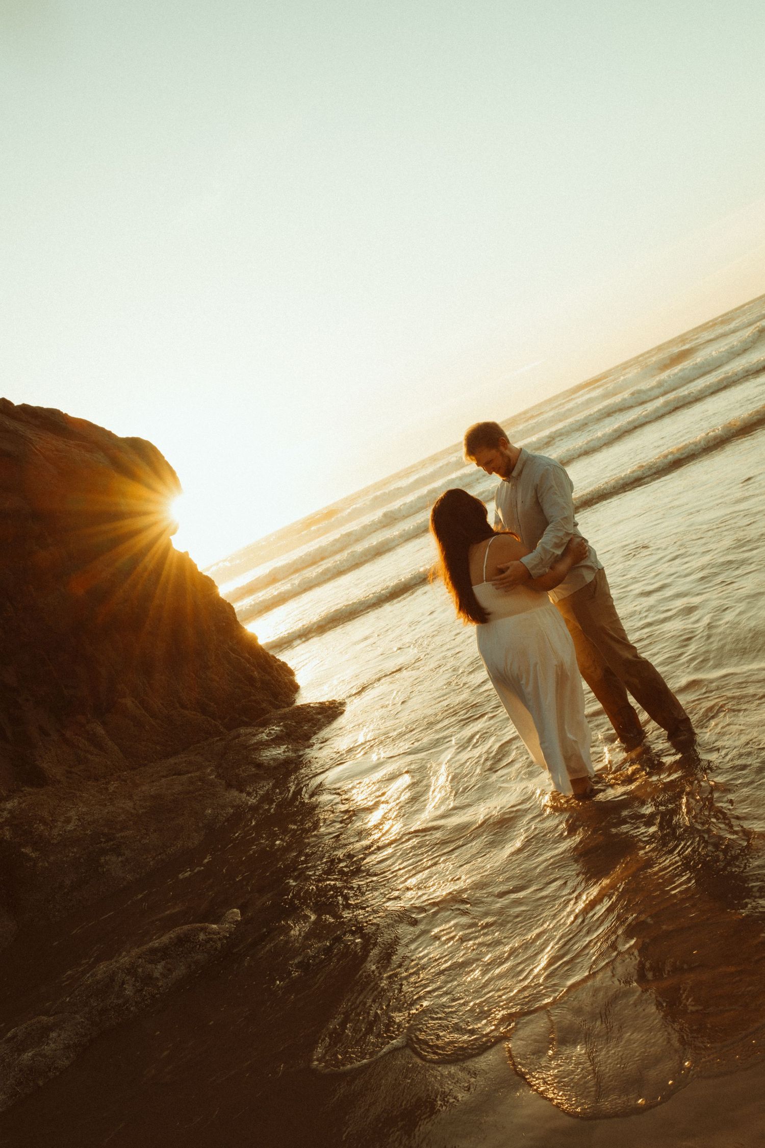 A couple shares a romantic moment on rocky coastal cliffs at sunset with golden light reflecting off the ocean waves.
