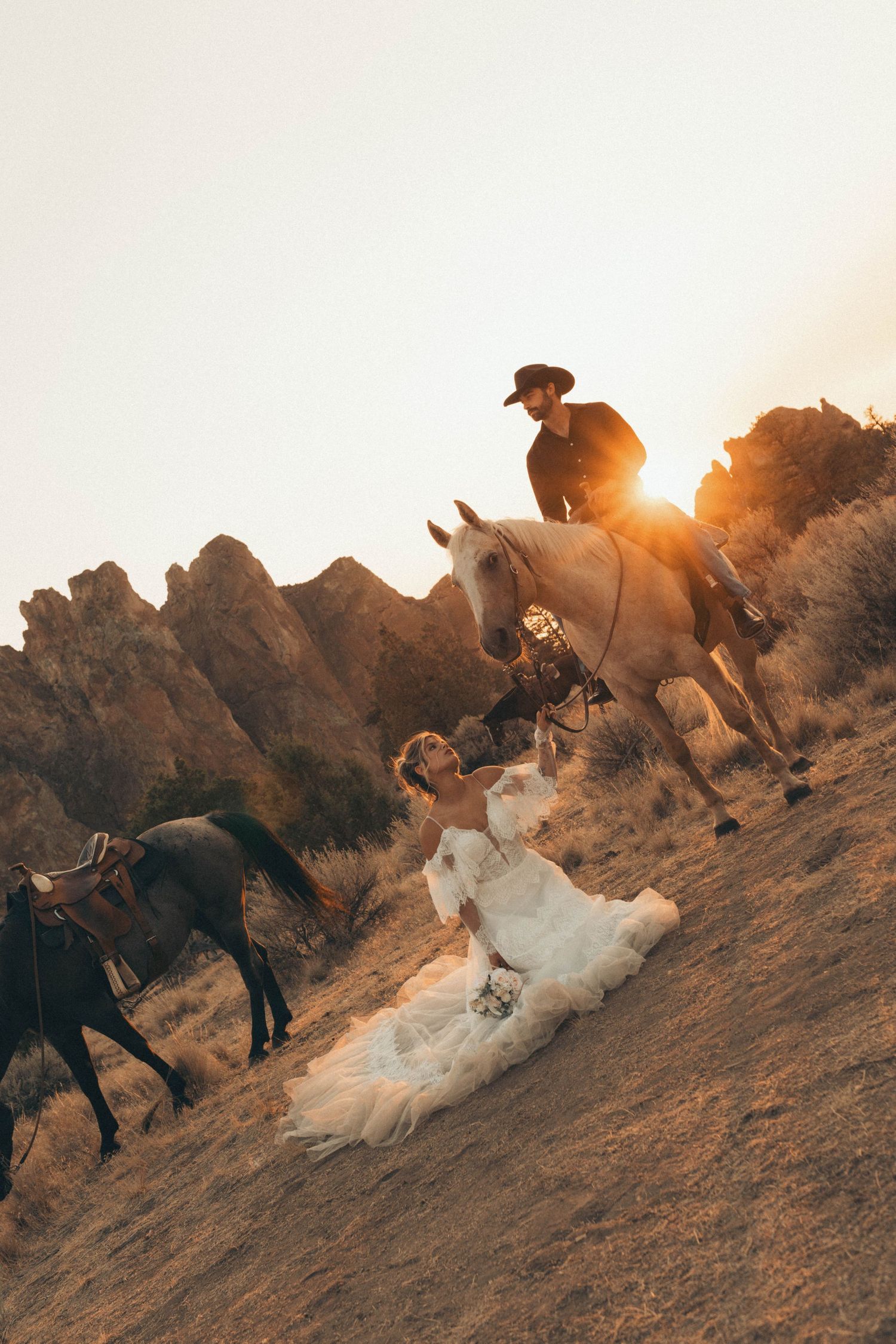 Bride in white gown sits on desert hillside as groom on horseback rides past rocky landscape at golden sunset.