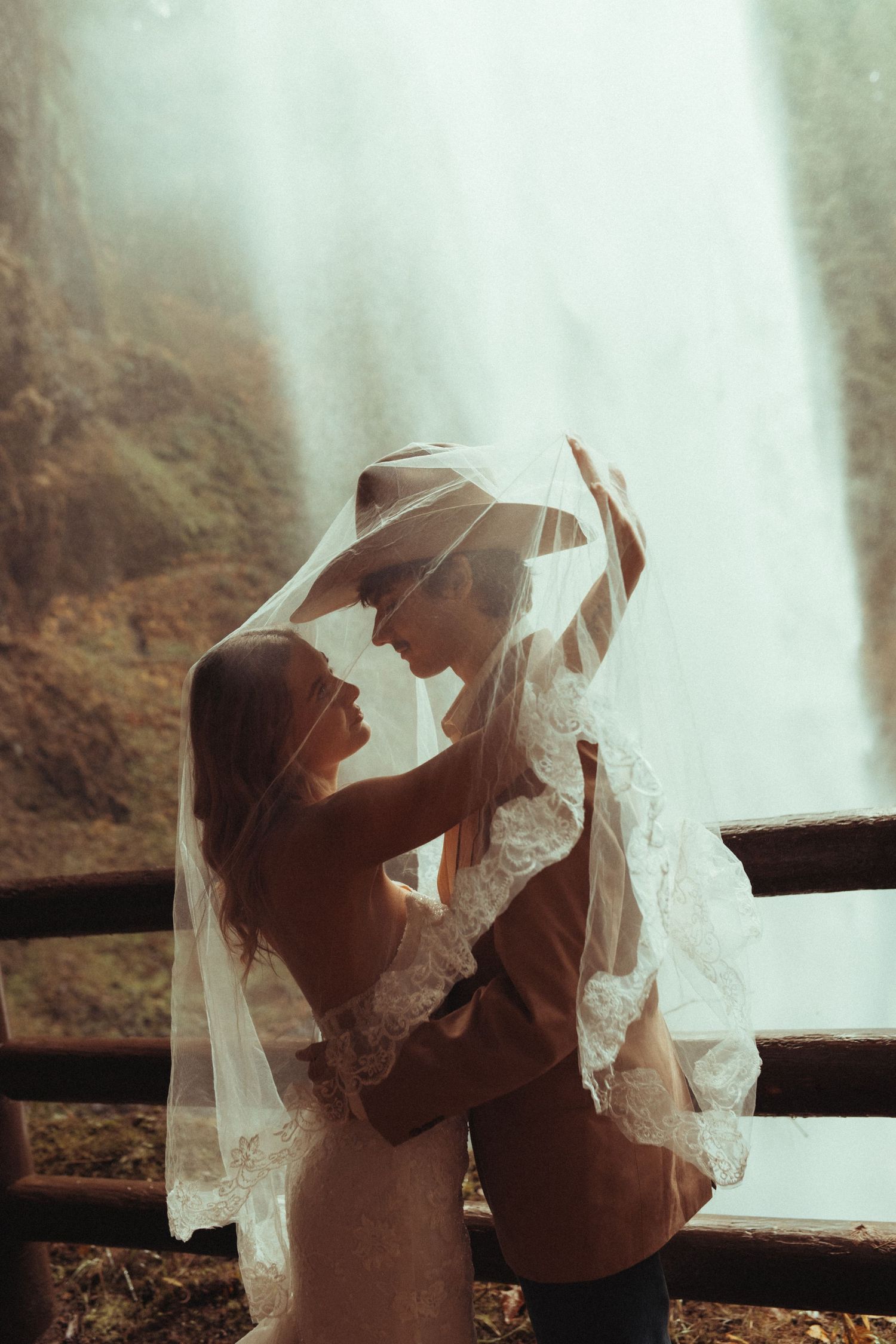 A romantic couple shares an intimate moment under a delicate lace veil against a misty waterfall backdrop.