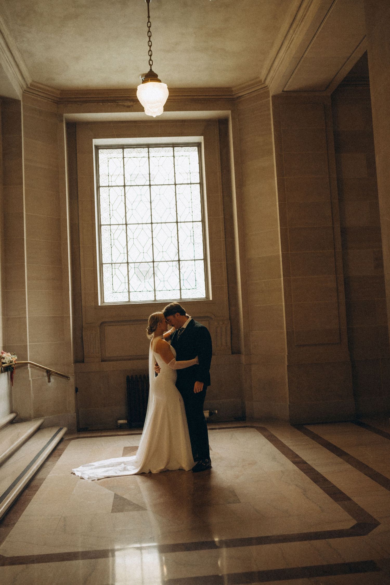 A romantic wedding couple embraces in front of a large window in an elegant hall with marble floors and vintage lighting.