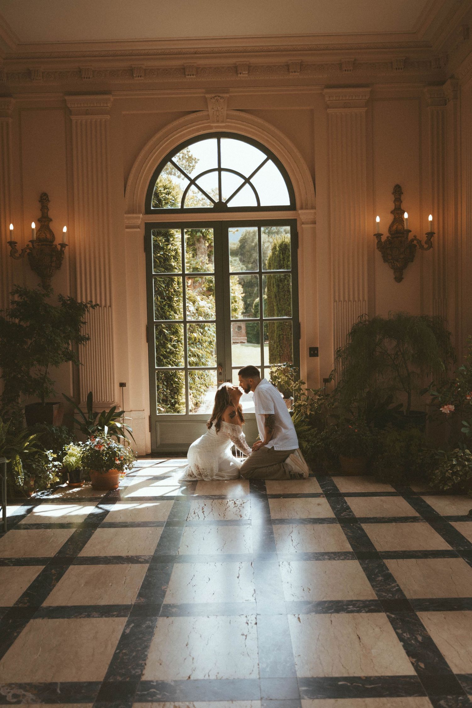 Couple in white kneeling together on a checkered marble floor in an elegant sunlit hall with an arched window and potted plants.