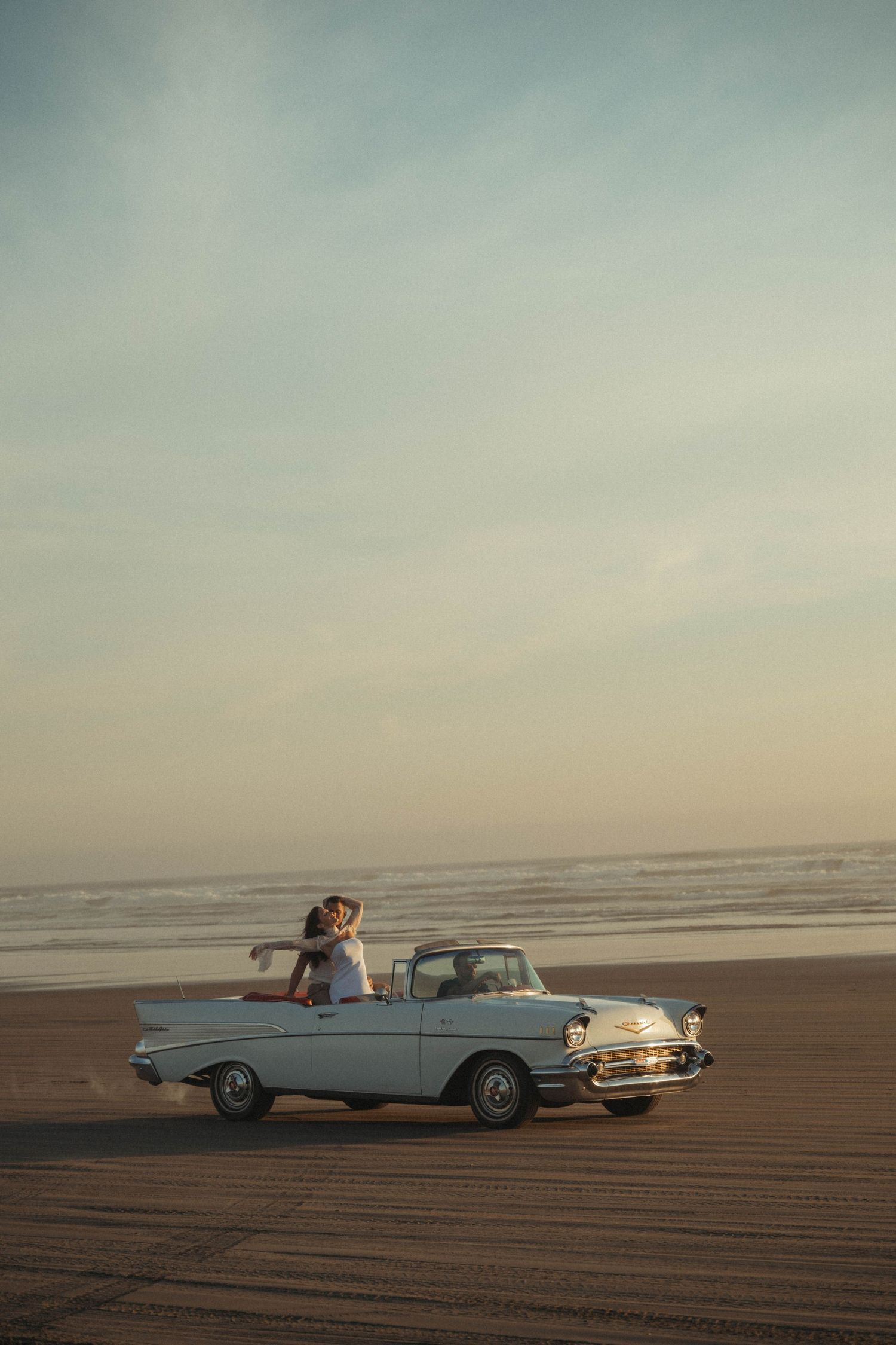 Couple embracing in a classic vintage convertible car parked on a sandy beach at sunset with ocean waves in the background.