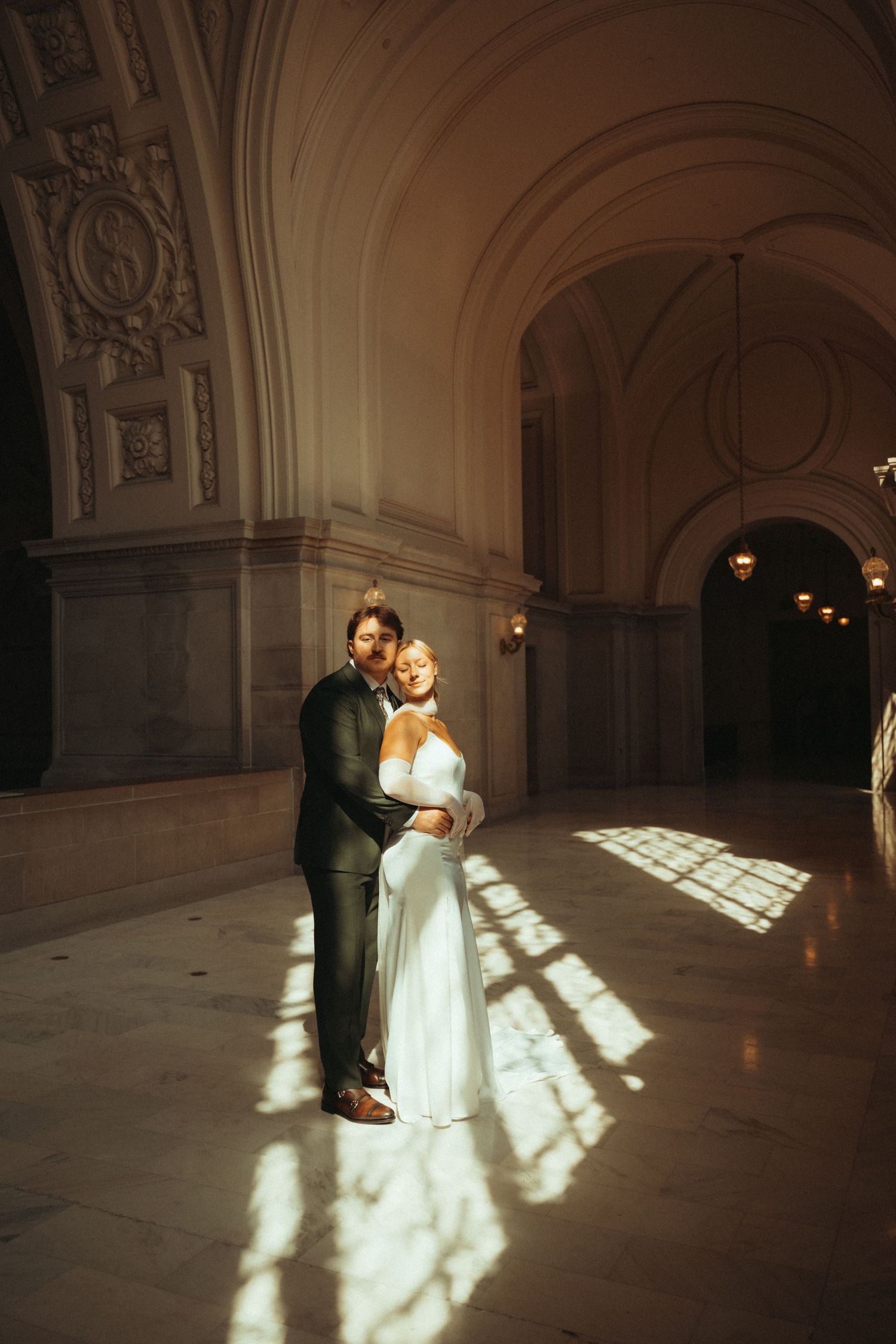 A couple embraces in an elegant hallway with ornate architecture as dramatic sunlight streams through windows creating patterns.
