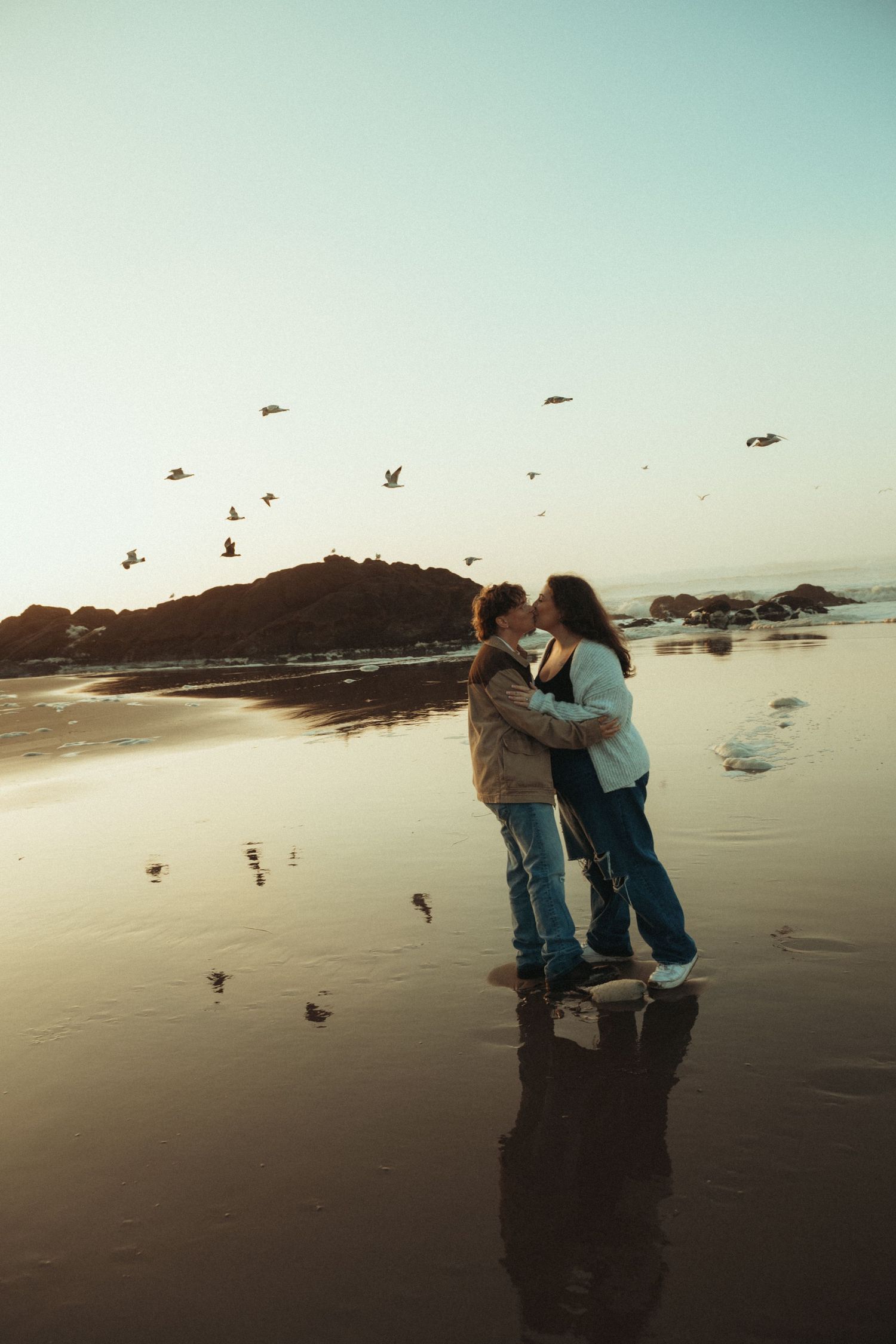Two people share a romantic moment on a misty beach at sunset with seabirds flying in the background.