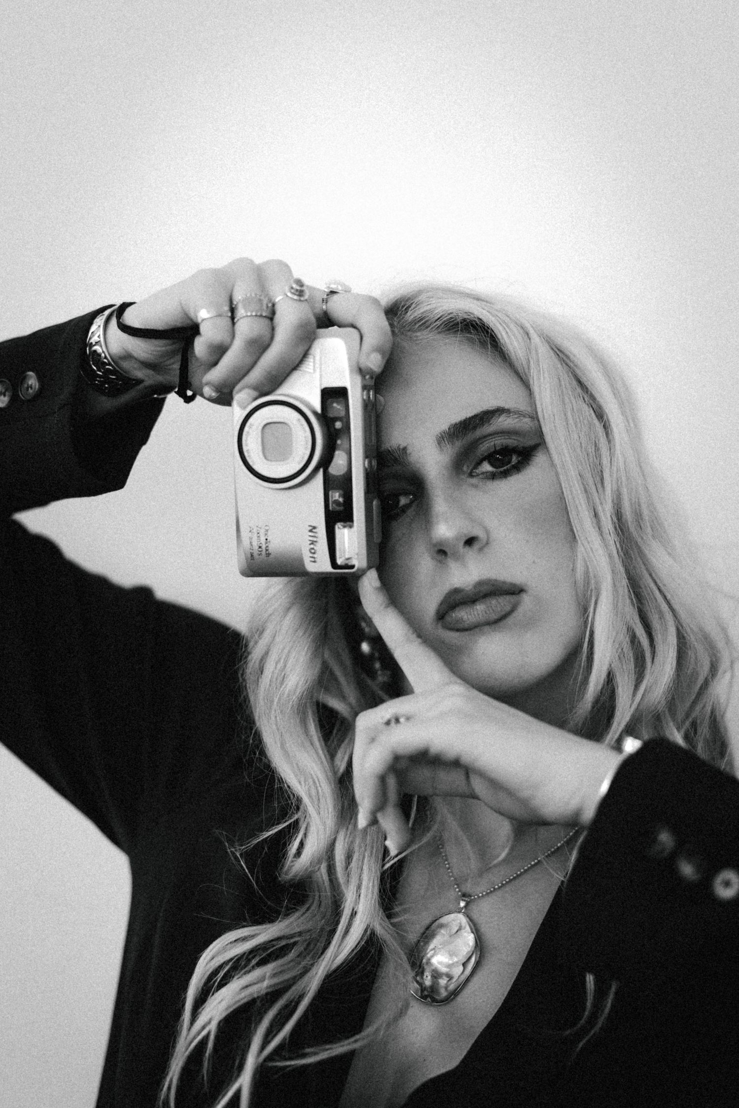 Black and white portrait of a woman with long wavy blonde hair holding a compact film camera to her face while wearing a pendant necklace.