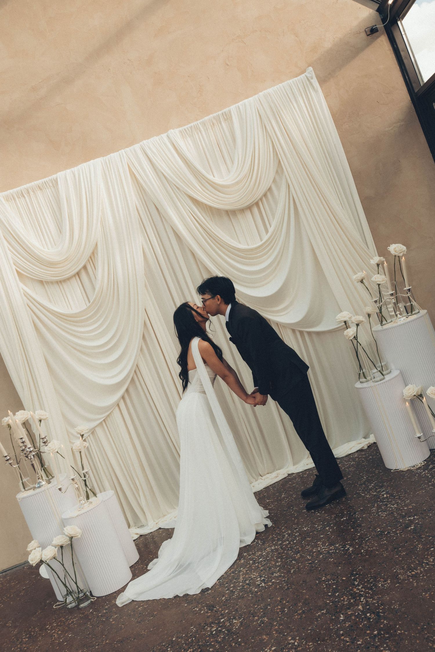 Bride and groom share a kiss at the altar in front of an elegant white draped backdrop with floral arrangements.