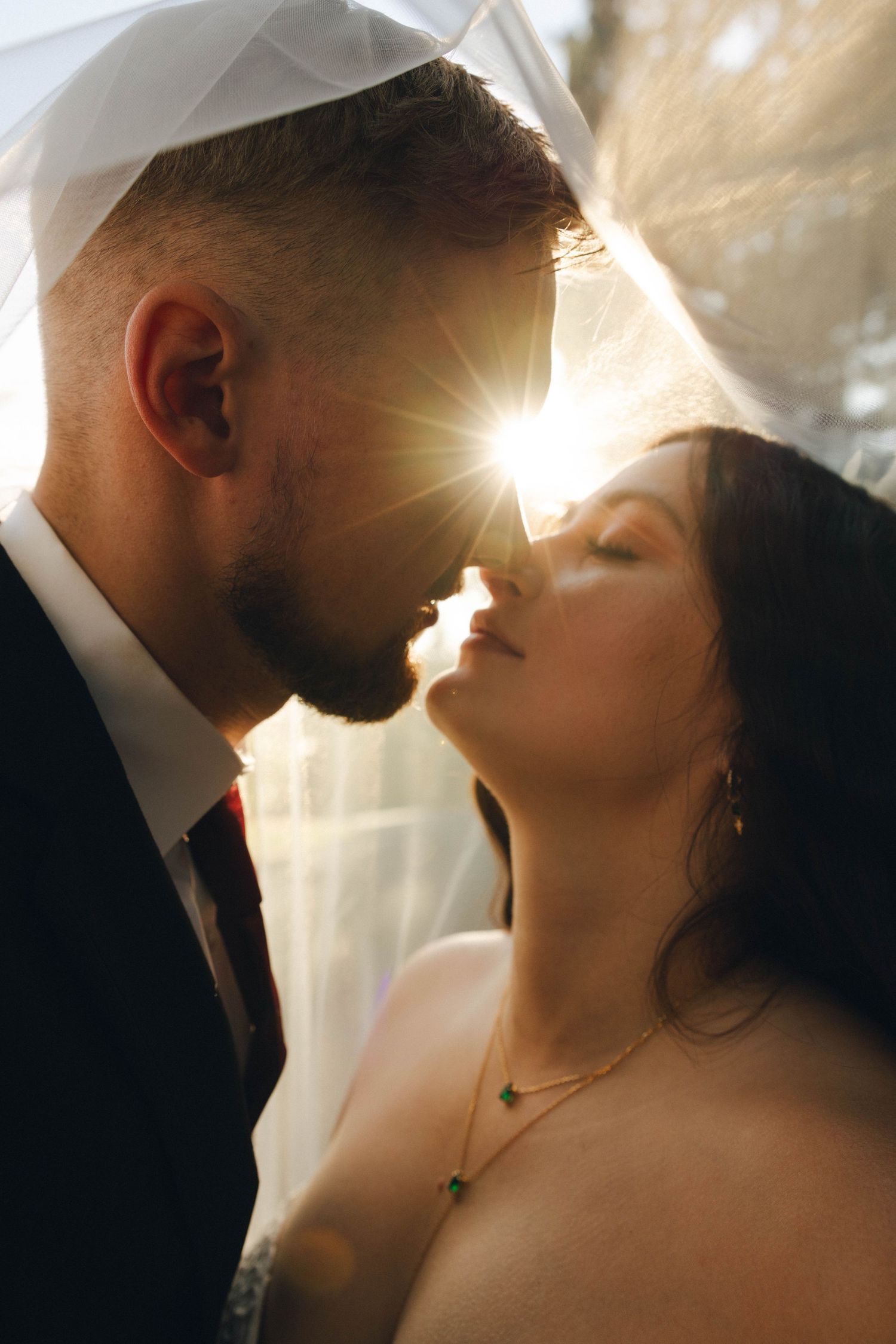 Bride and groom share romantic moment under veil with golden sunlight glowing between them at wedding.
