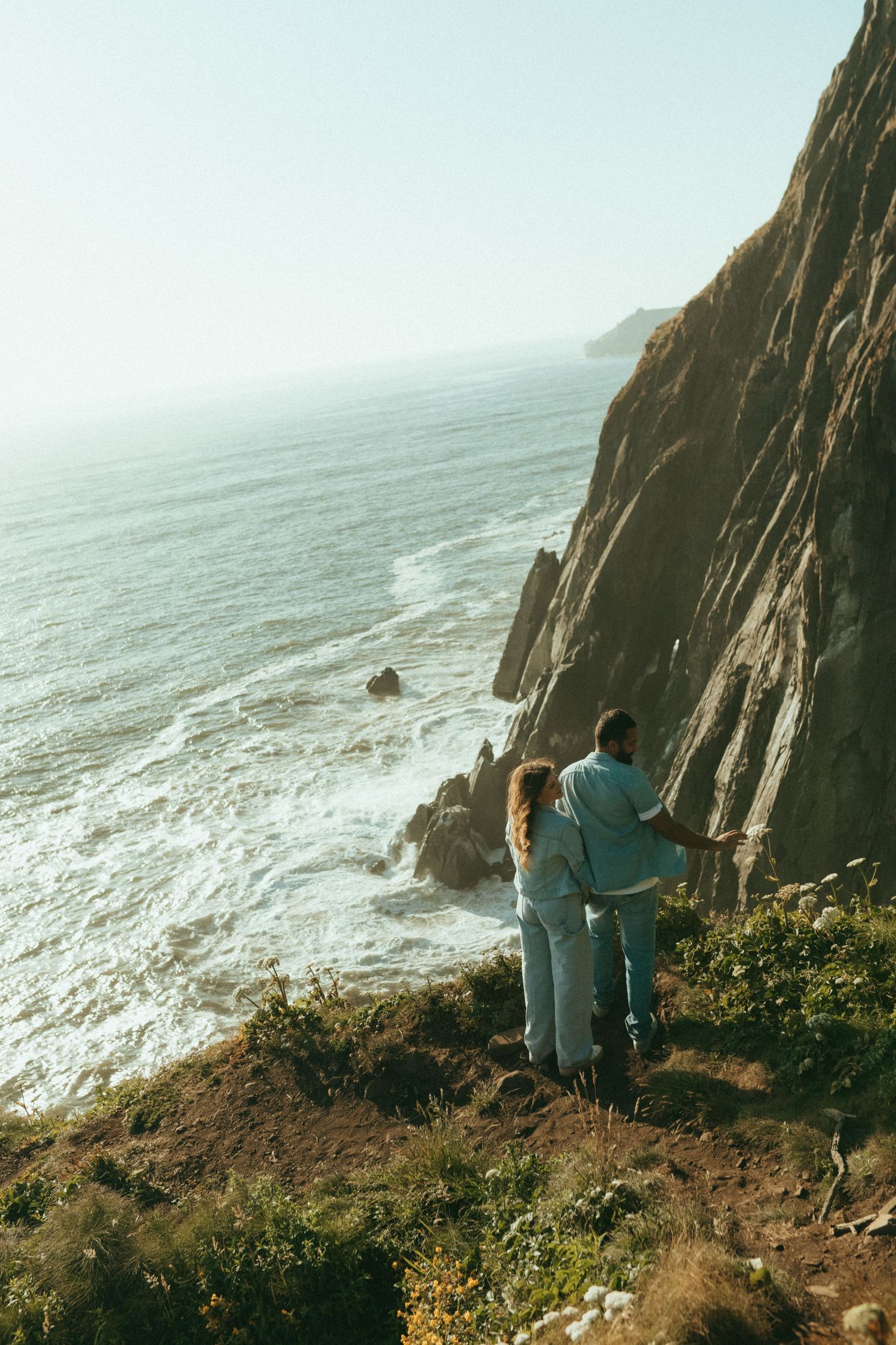 Couple standing at edge of coastal cliffs overlooking dramatic ocean views along rugged Pacific coastline.