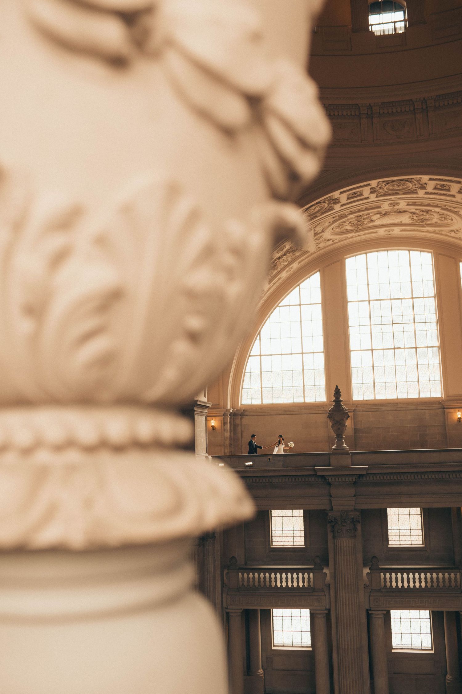 Ornate carved stone column in the foreground with a couple standing on a balcony beneath grand arched windows in a historic domed building.