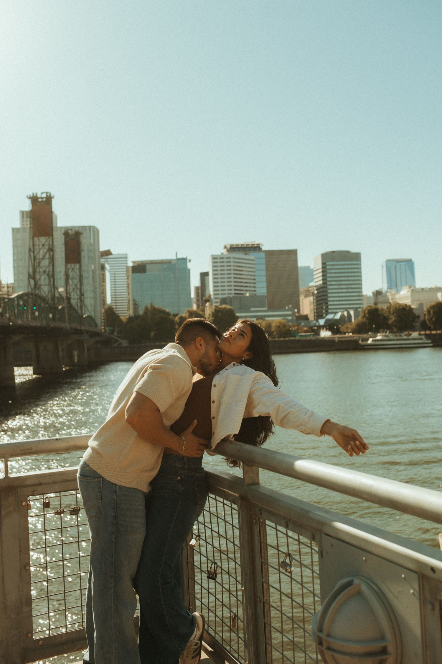 A couple leans over a waterfront railing overlooking a cityscape with tall buildings and sparkling water.