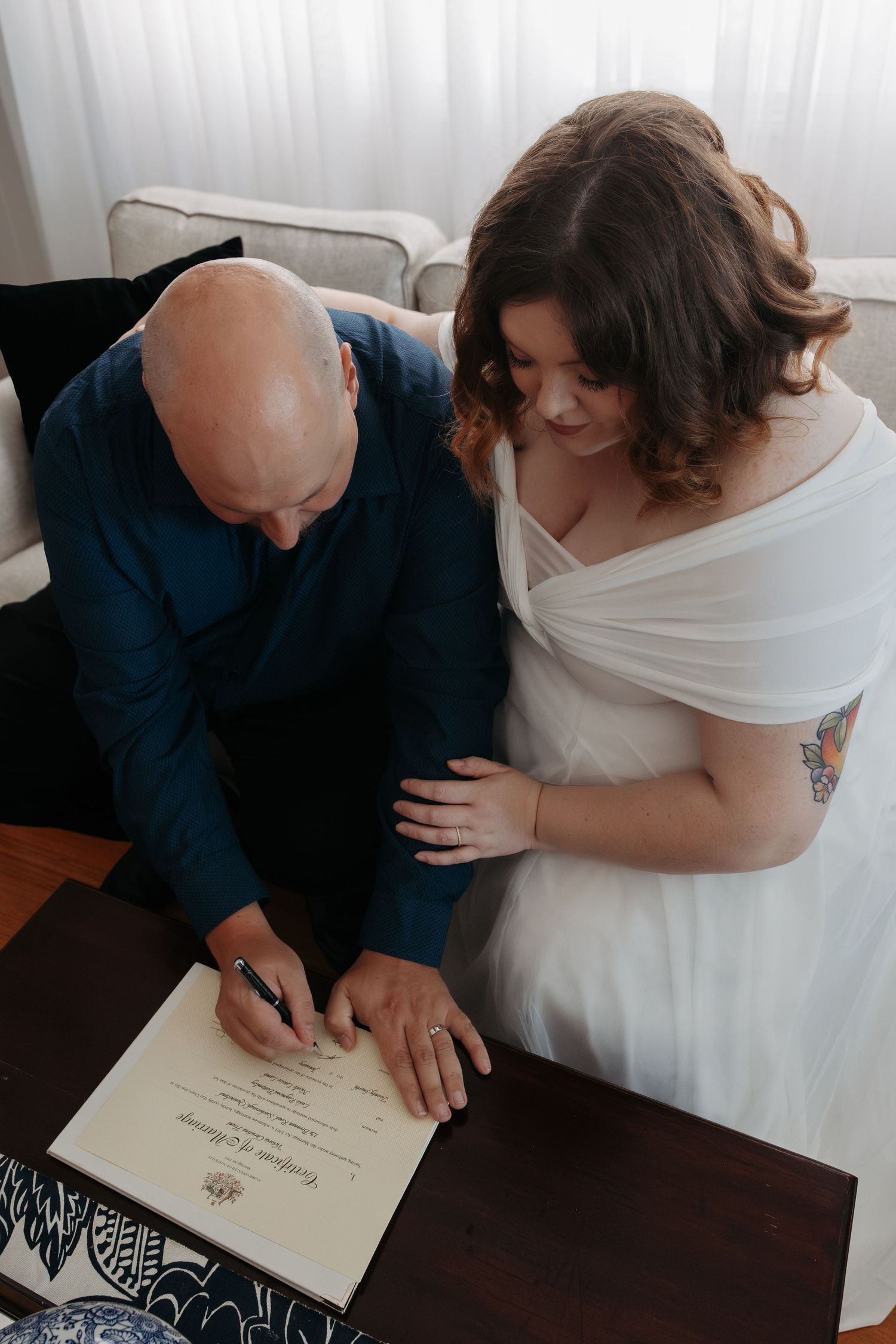 A couple signs their marriage certificate at a dark wooden table during an intimate indoor ceremony.