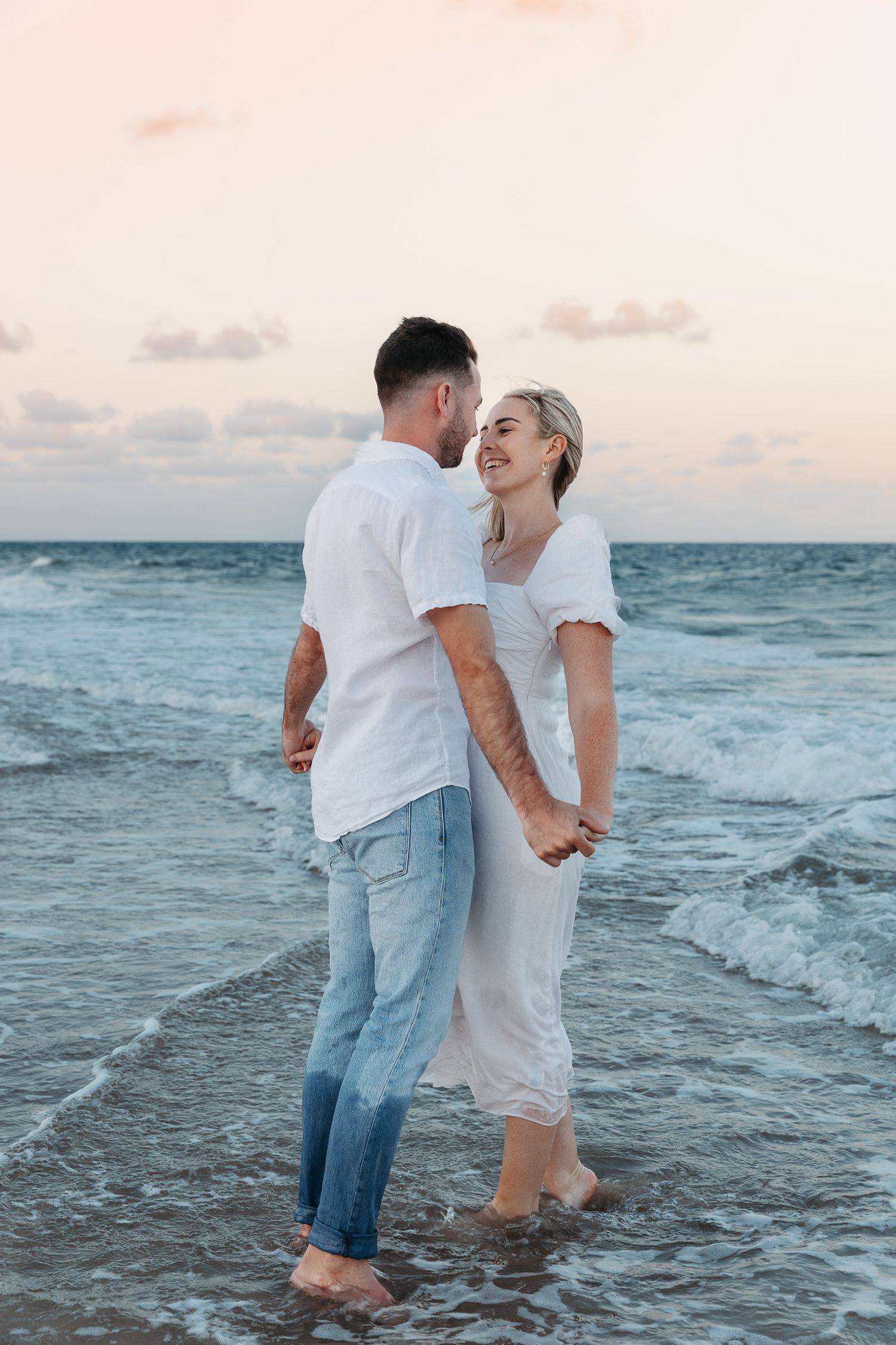 A romantic couple stands barefoot in the ocean waves at sunset wearing coordinated white outfits.