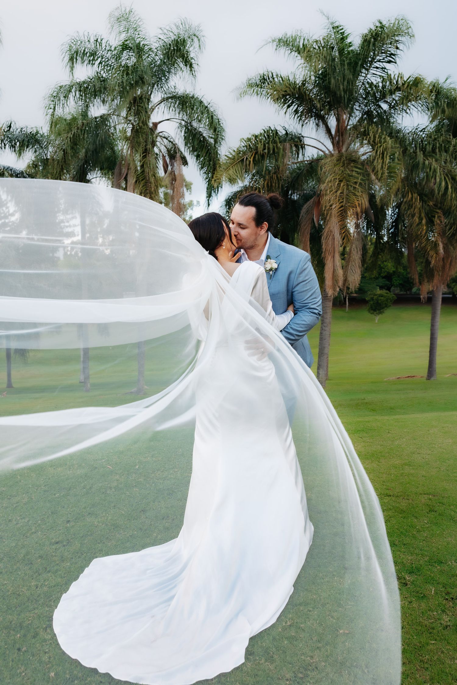 A romantic wedding couple embraces on a green lawn surrounded by palm trees as the bride's long veil flows in the breeze.