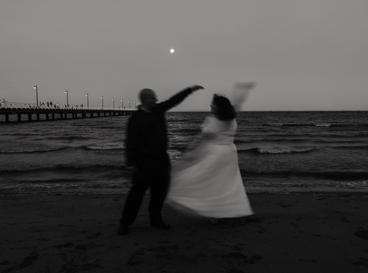 Silhouetted couple dancing on a beach at dusk with a pier and moon in the background.