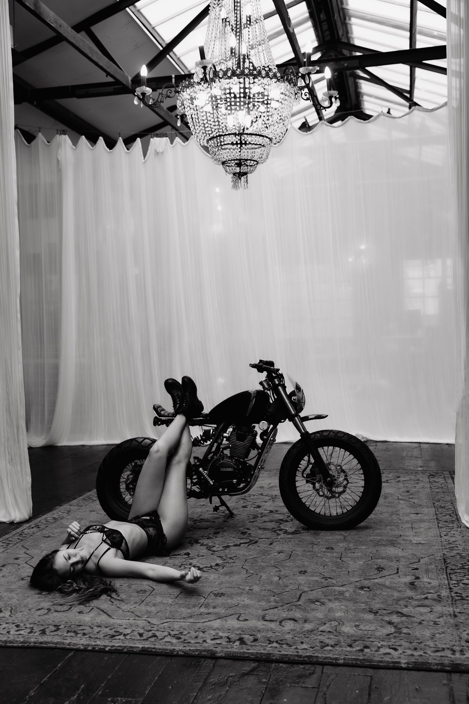 Black and white artistic scene of a motorcycle beneath a grand chandelier with flowing white curtains.