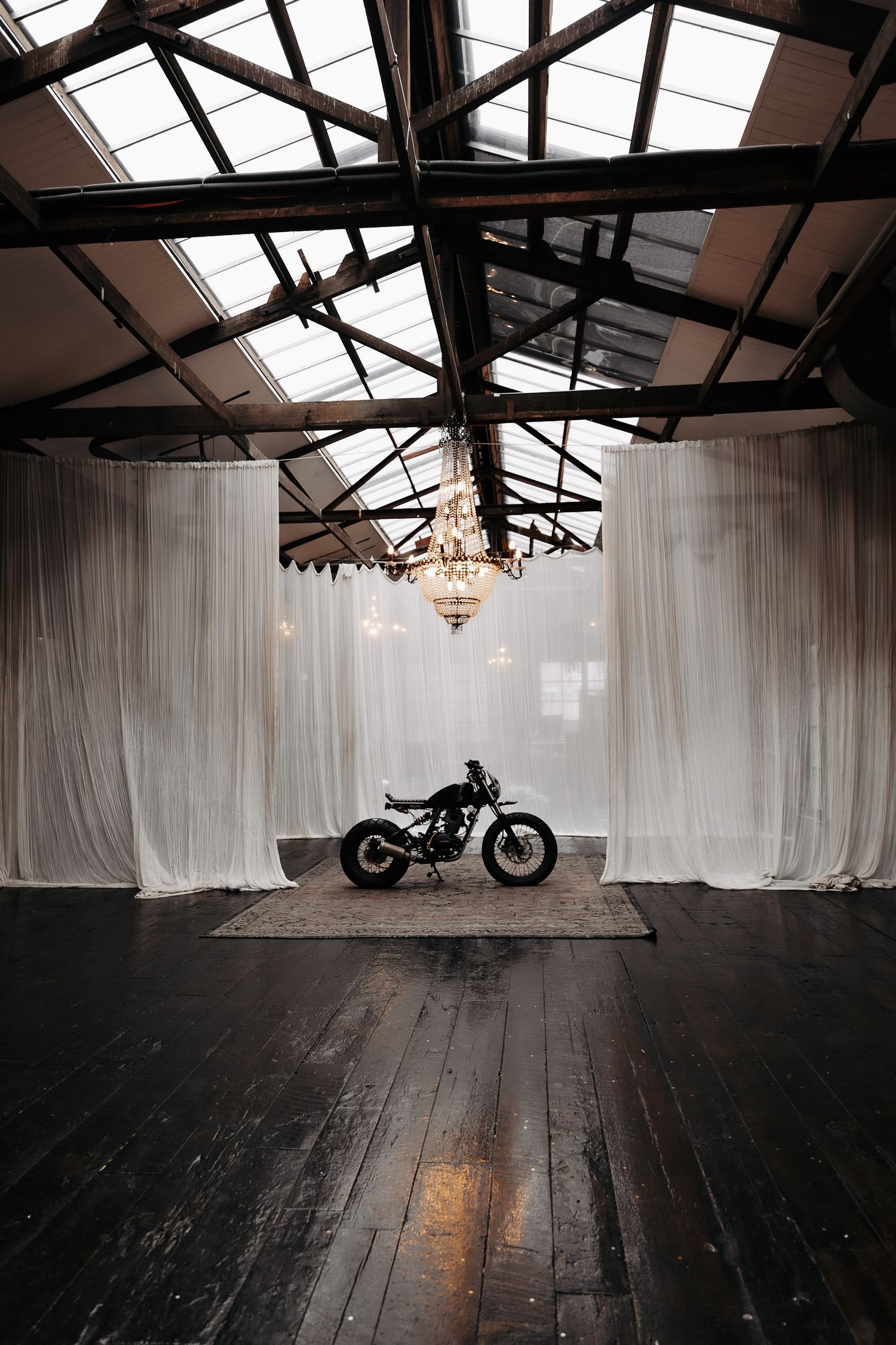 Vintage motorcycle silhouetted against white curtains in an industrial space with exposed wooden beams and a chandelier.