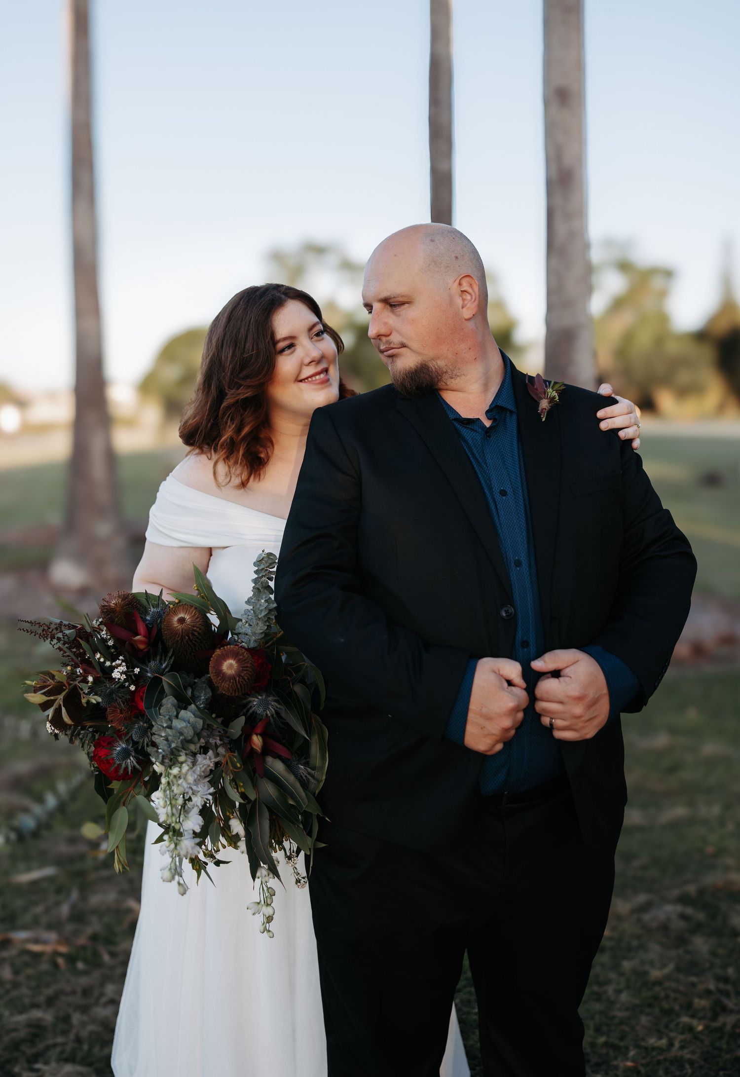 A romantic wedding couple embraces outdoors with palm trees and a beautiful burgundy and white floral bouquet.