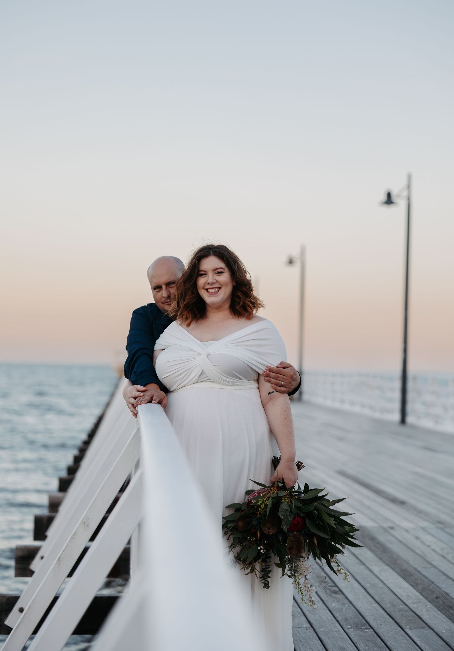 A couple shares an intimate moment on a wooden pier at sunset while holding a green floral bouquet.