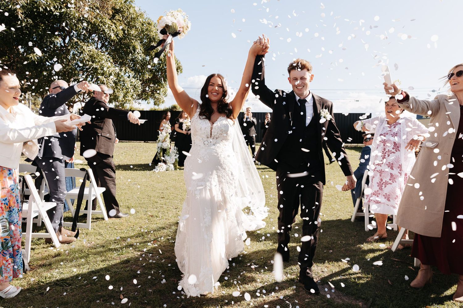 Wedding guests joyfully toss white flower petals as newlyweds walk through during an outdoor ceremony celebration.
