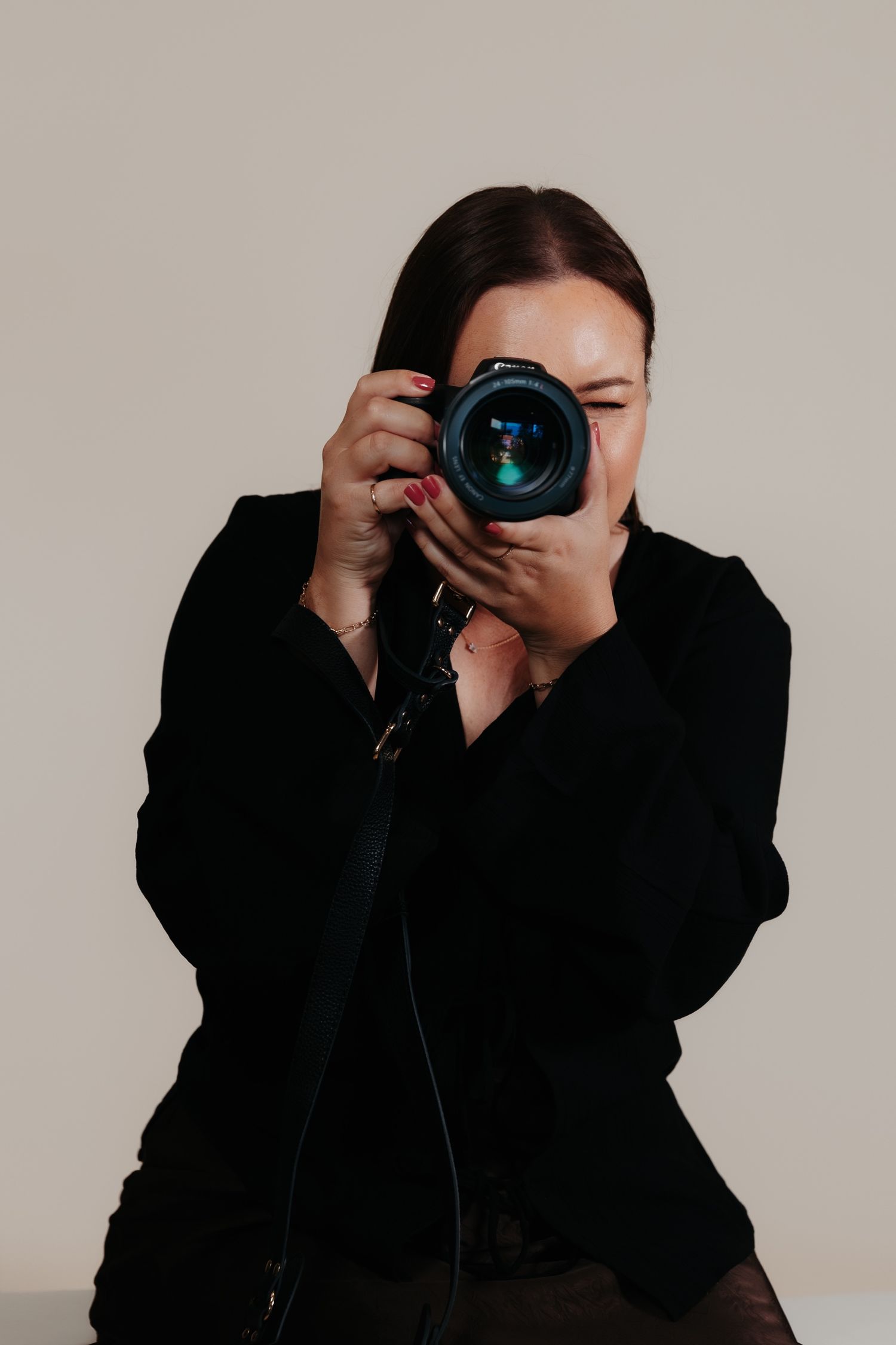 Professional photographer wearing black clothing holds a DSLR camera with large lens against beige background.