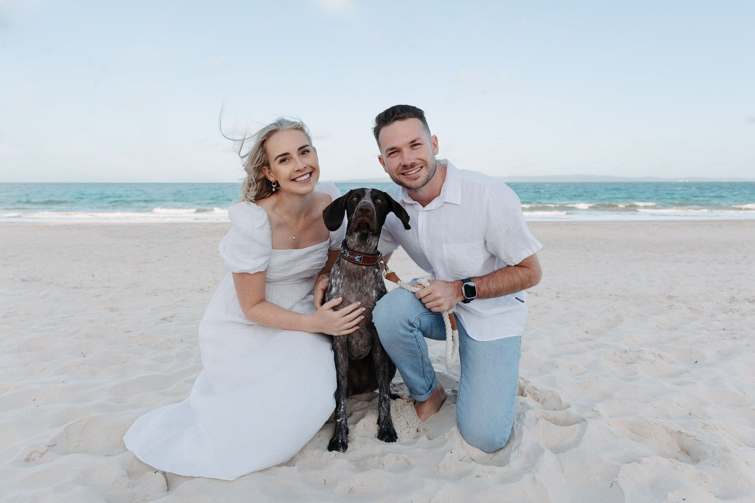 A couple poses on a sunny beach with their large black dog, dressed in casual white attire against an ocean backdrop.