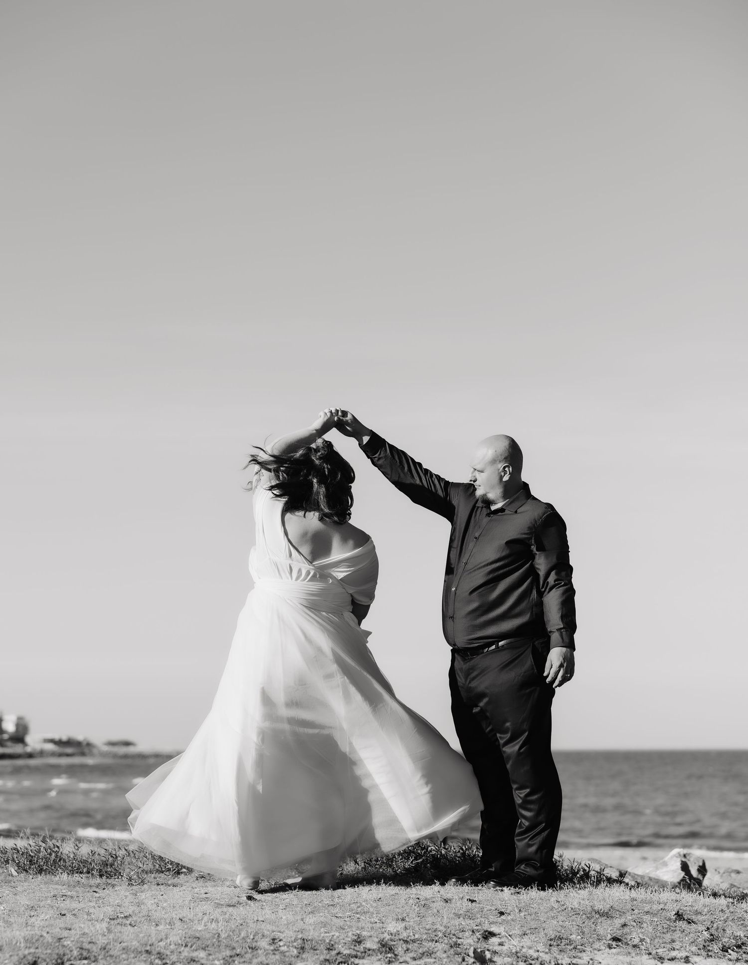 A joyful couple dances together on a coastal cliff in black and white, with the bride's white dress flowing in the breeze.