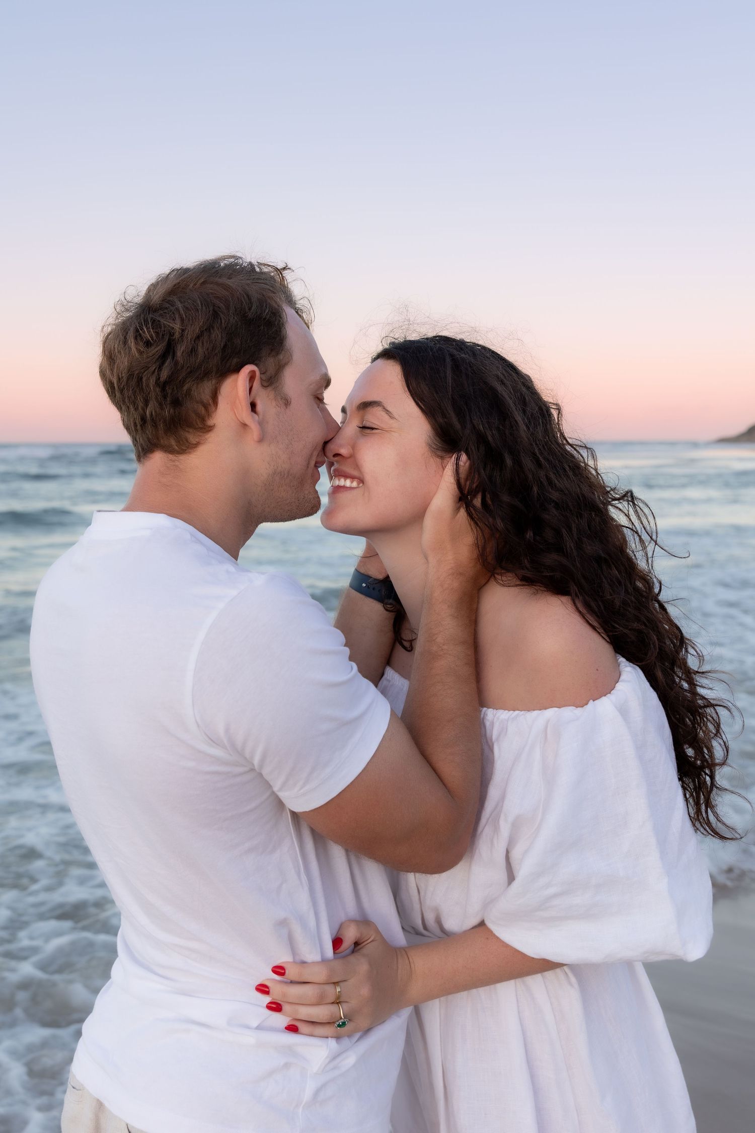 Romantic couple in white outfits embrace on beach at sunset with ocean waves in background.