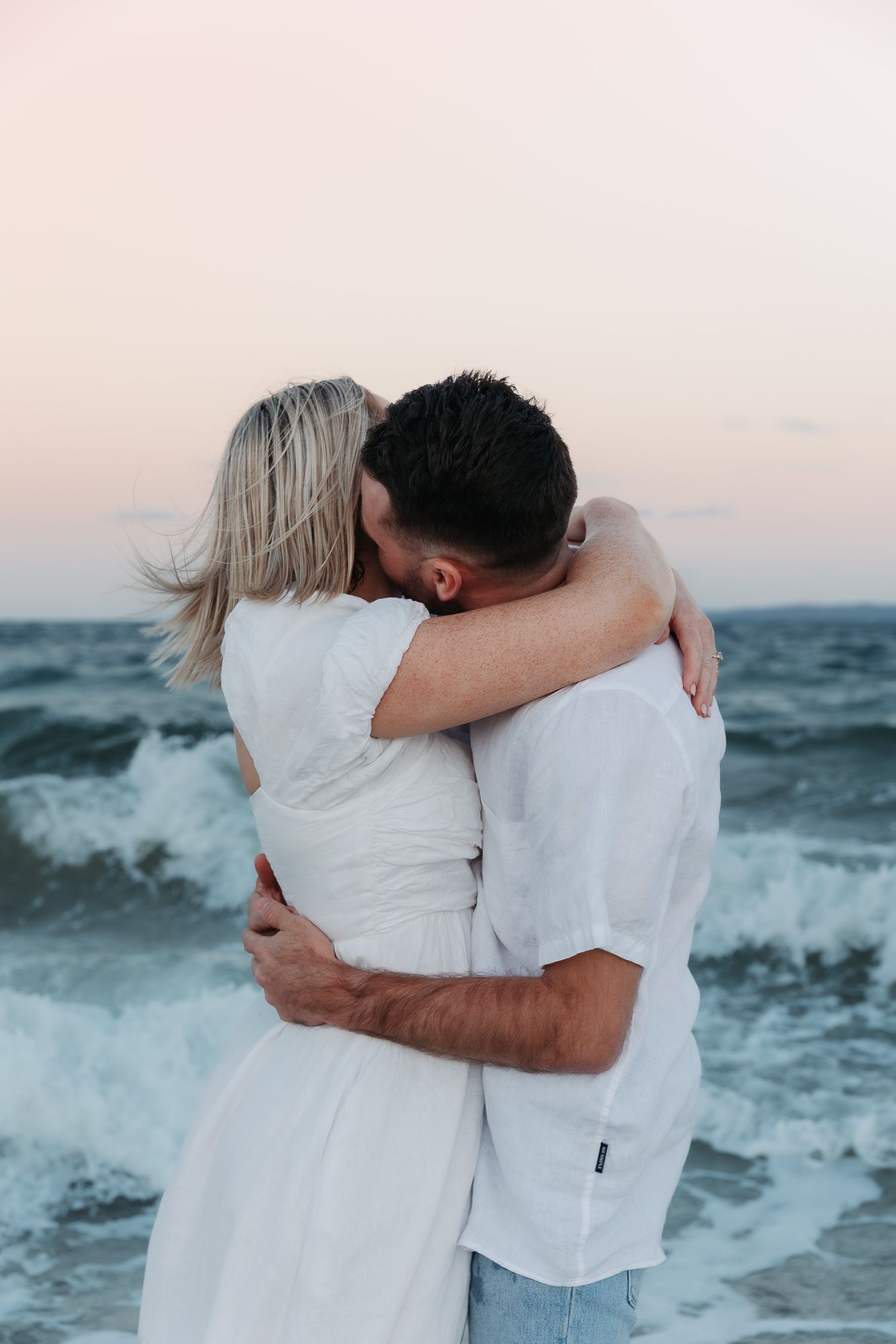 Couple in white clothing embrace while standing by crashing ocean waves at sunset.