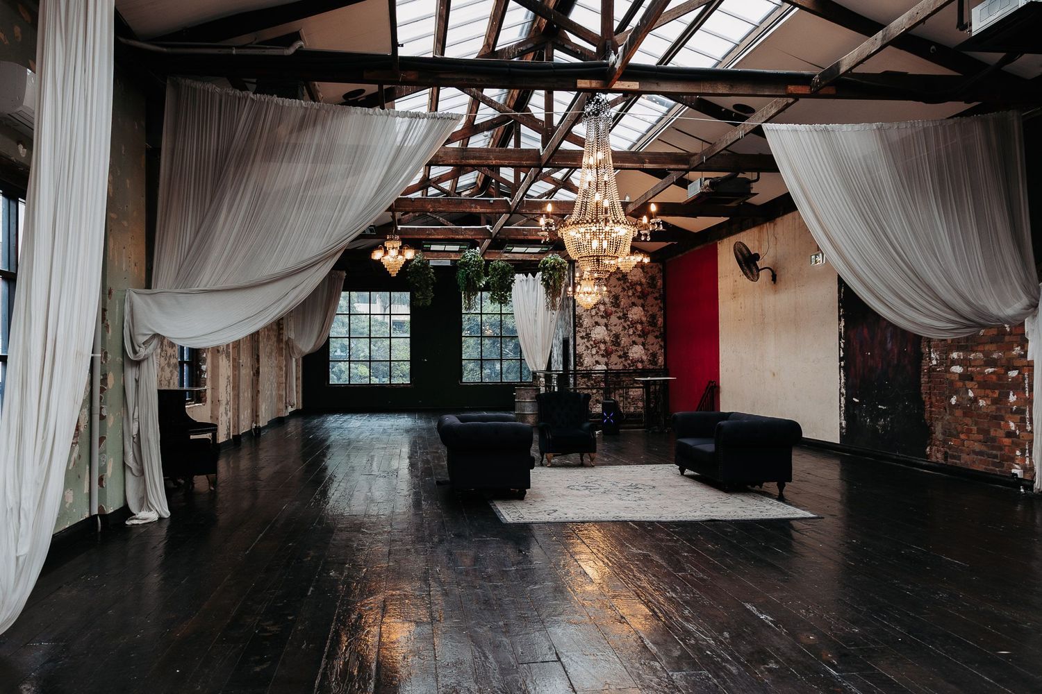 Dark rustic event space with exposed wooden beams, draping white curtains, a chandelier and hardwood floors.